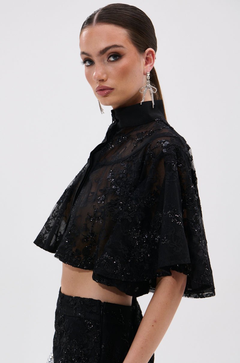 A woman with slicked-back hair and bold makeup wears the QUEEN B LACE BUTTON DOWN, featuring sheer black embroidered fabric with sequin details. She pairs it with a matching skirt and statement earrings, posing against a light background.