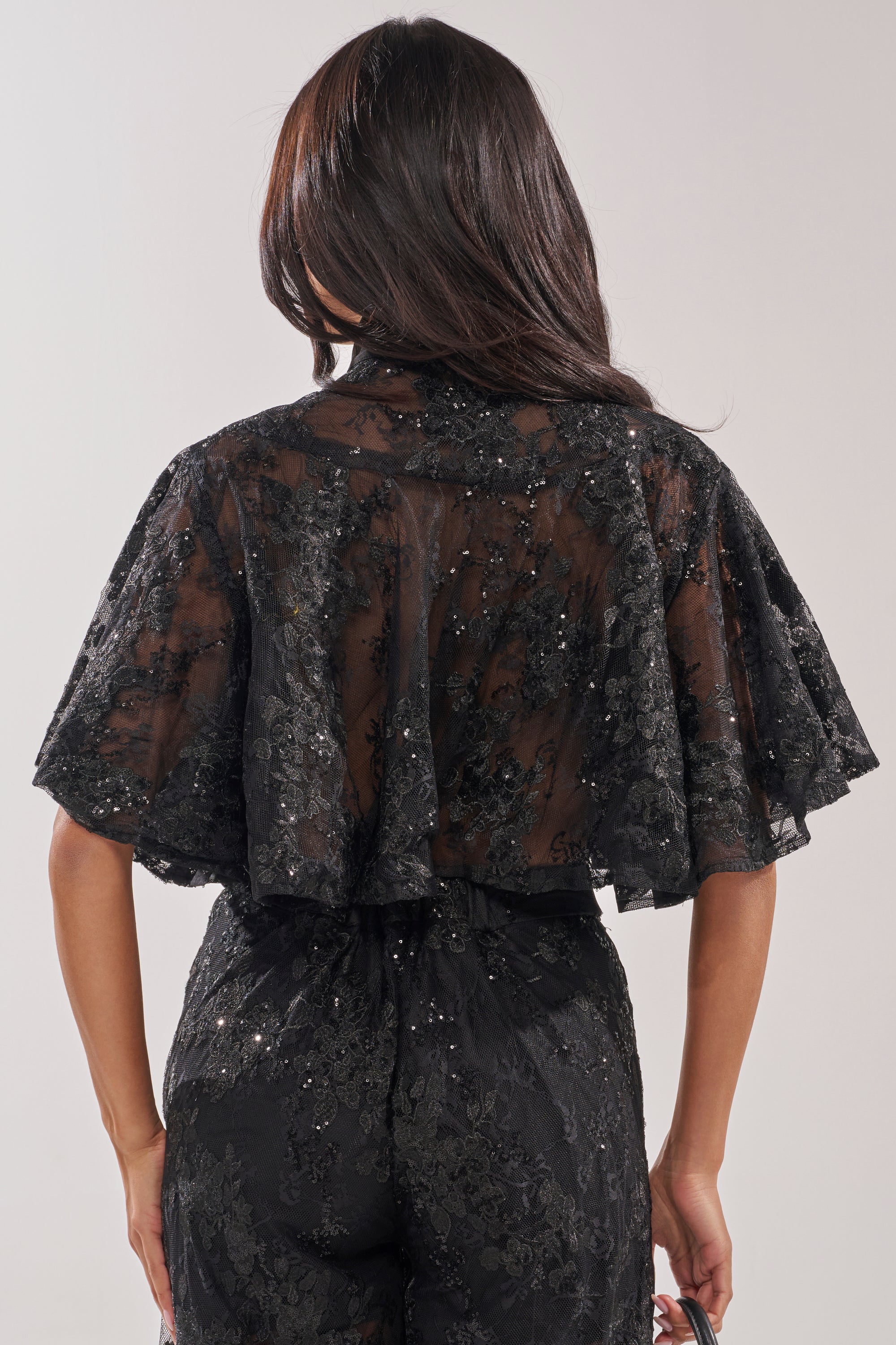 A woman with long, dark hair faces away, wearing the QUEEN B LACE BUTTON DOWN—a sheer black lace top with floral patterns and short, flared sleeves—paired with matching high-waisted black lace pants. 3