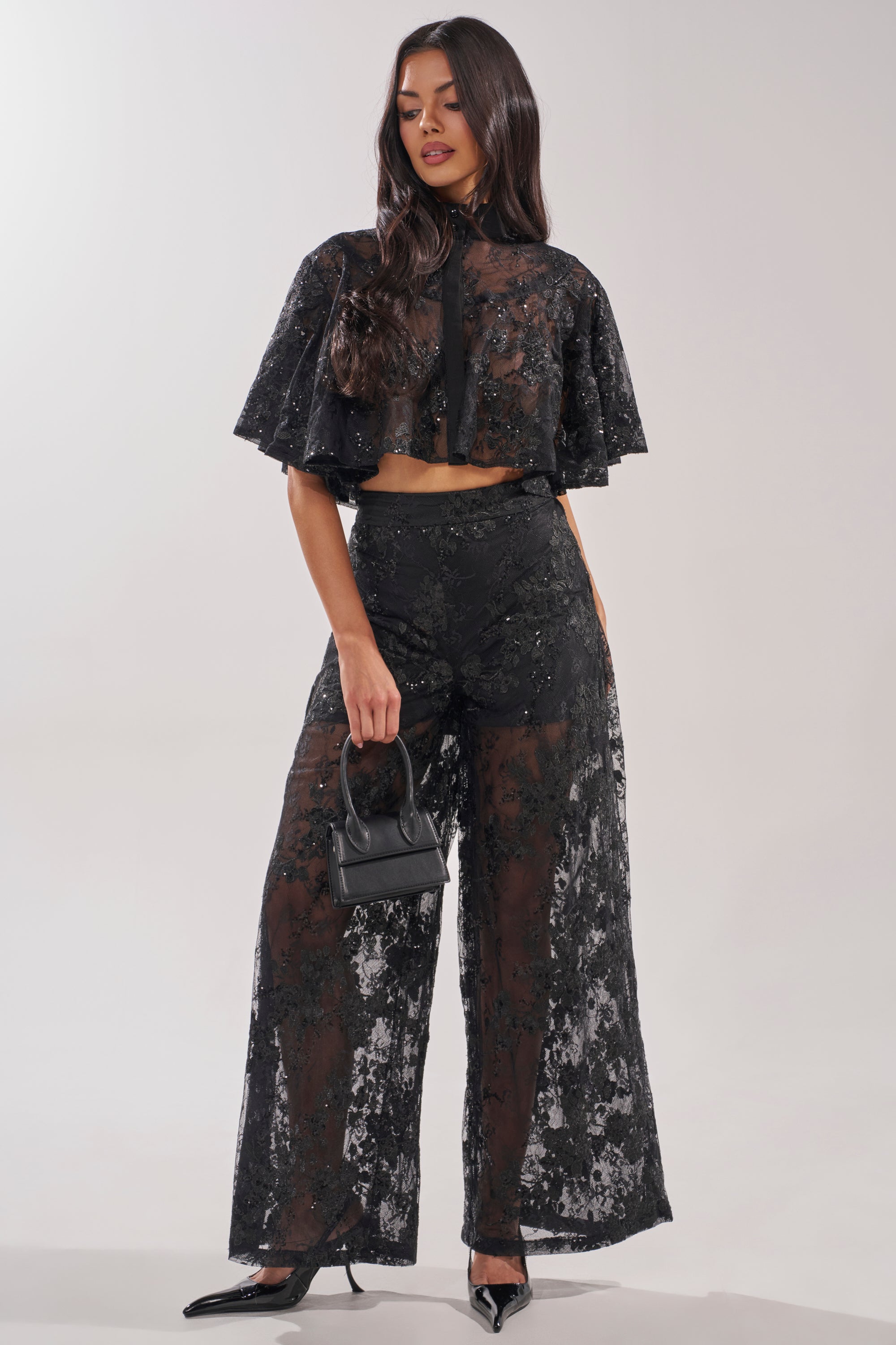 A woman poses in the QUEEN B LACE BUTTON DOWN, a sheer floral black lace top with wide-leg pants. She accessorizes with a small black handbag and pointed black heels. 4