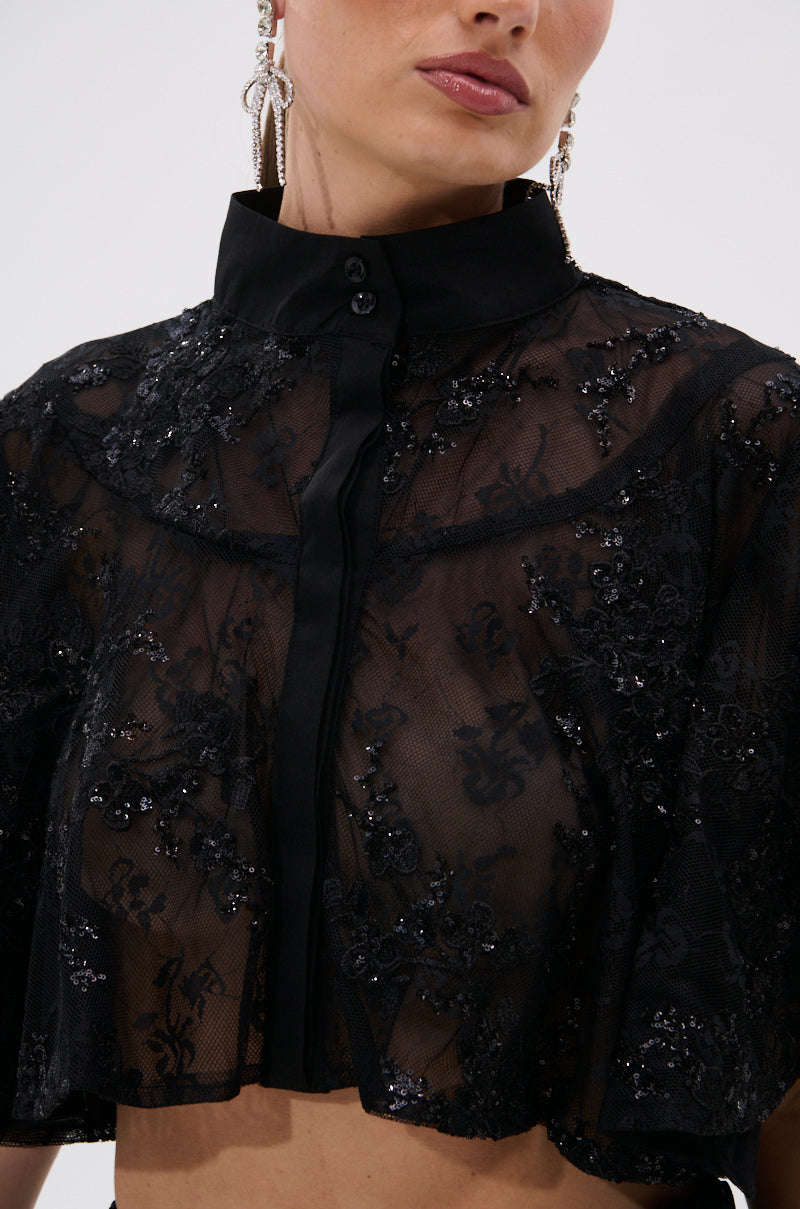 A woman wears the QUEEN B LACE BUTTON DOWN, a sheer floral mesh top with intricate embroidery, sequin details, high collar, and statement earrings.