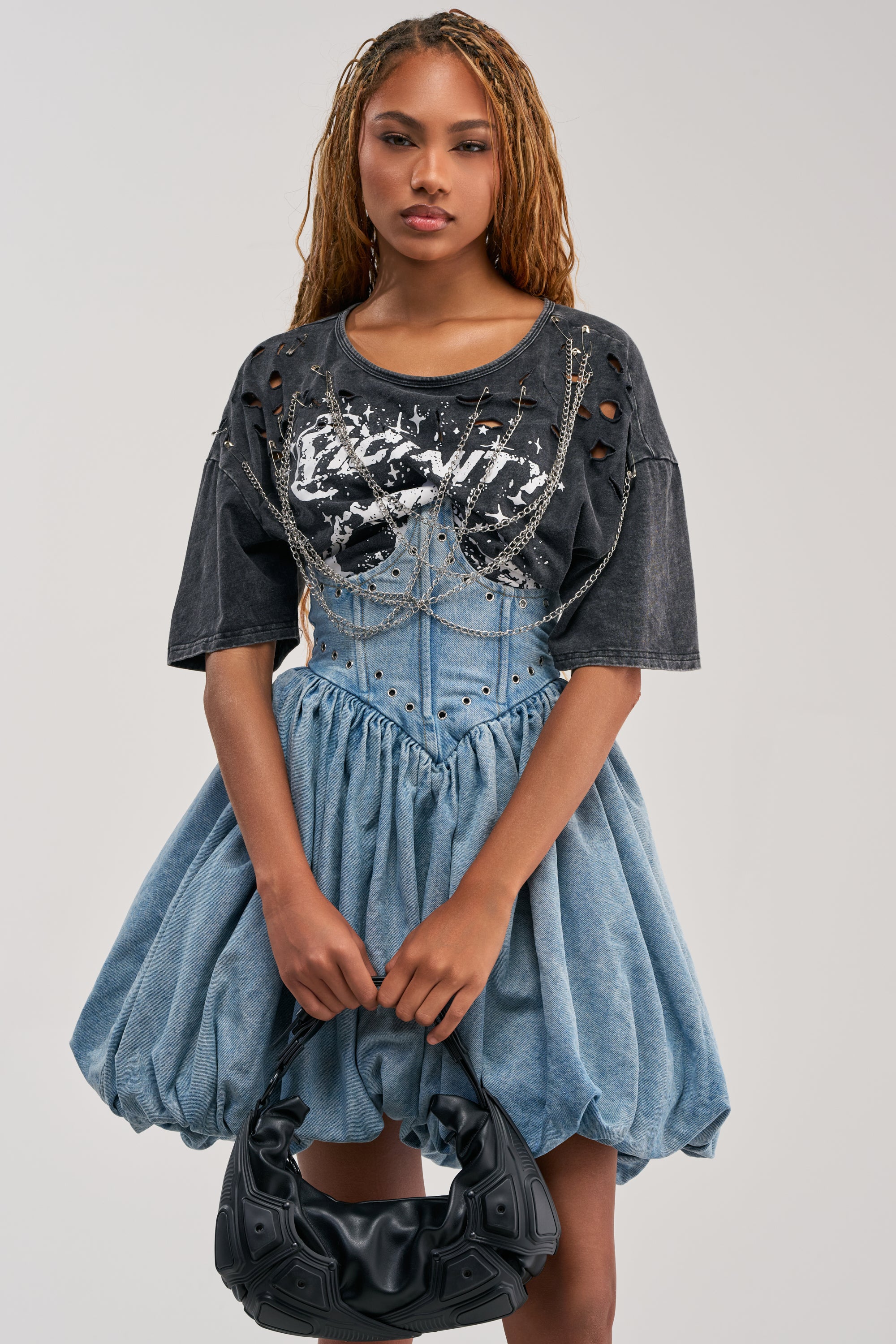 A woman with long braids wears the COOL GIRLS ONLY MINI DRESS—a voluminous blue denim bubble dress—layered under a ripped black graphic t-shirt and paired with a black textured handbag. Her look captures cool streetwear vibes as she poses against a plain light background.