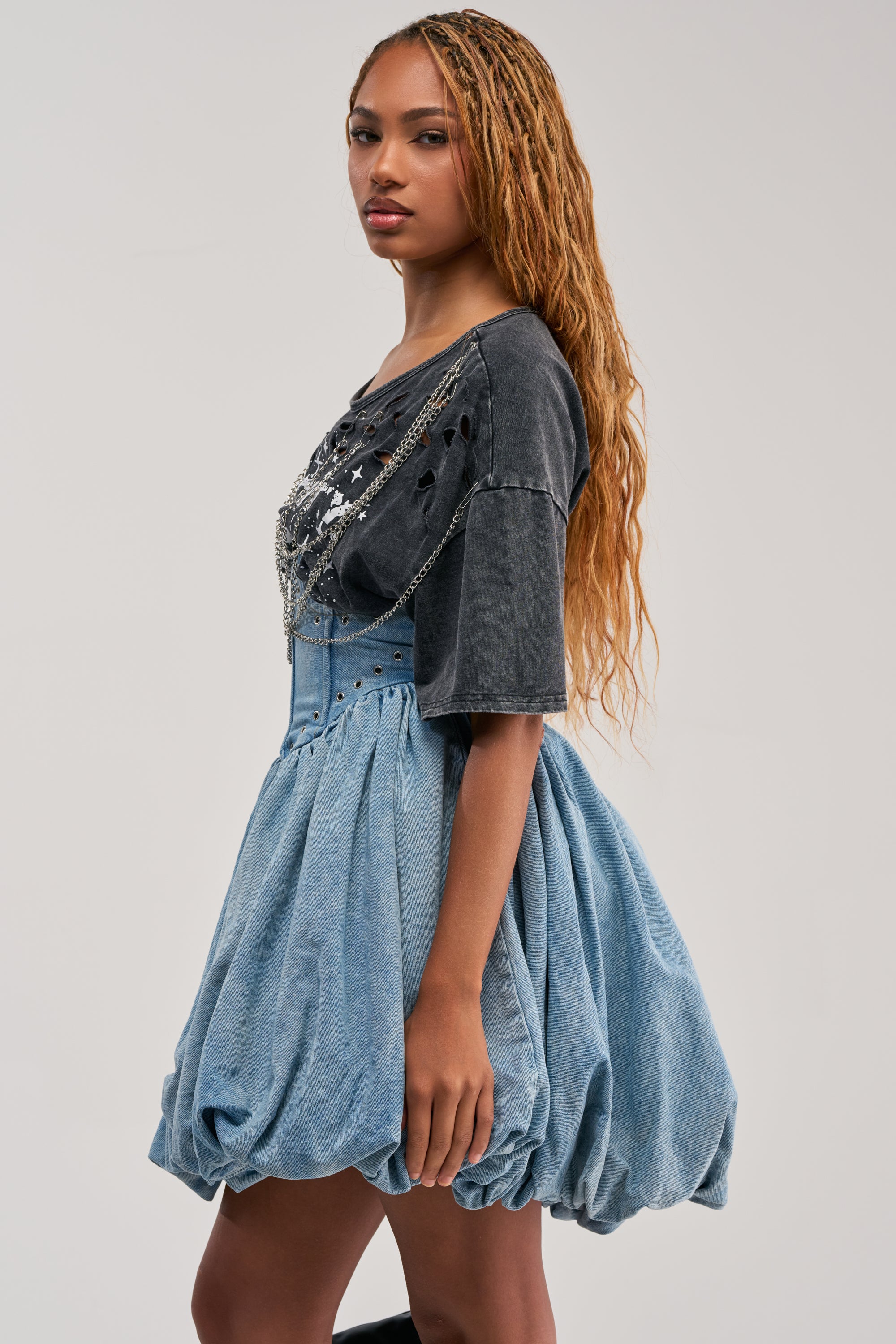 Wearing the COOL GIRLS ONLY MINI DRESS, a woman with long, wavy hair stands sideways against a plain light background. She styles the look with streetwear flair and gazes at the camera, showing off the dress’s high-waisted, voluminous blue skirt and distressed black top.