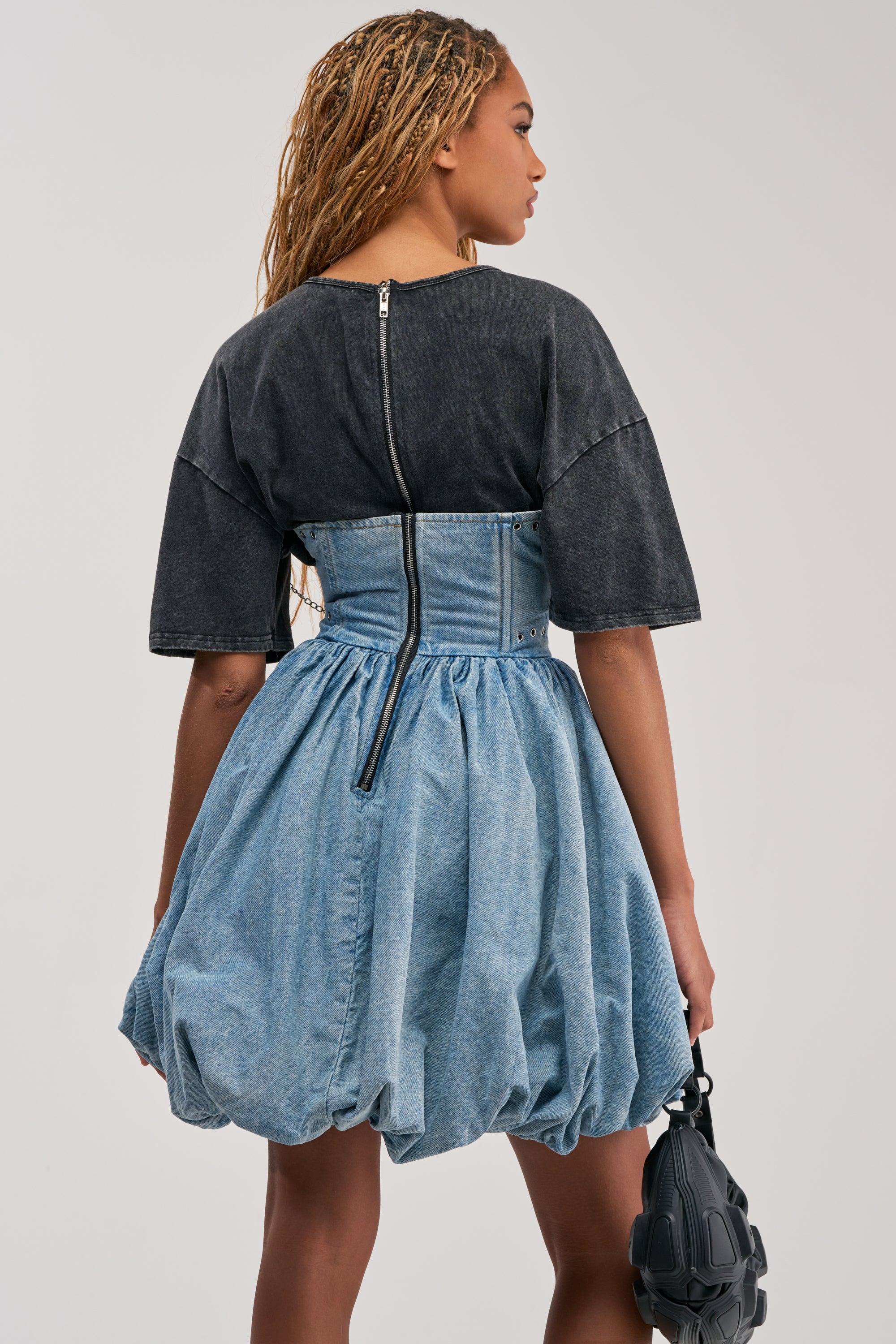 A person with braided hair is seen from behind, showcasing streetwear styling in the black COOL GIRLS ONLY MINI DRESS with short sleeves and a zipper detail, paired with a blue denim bubble skirt. They hold a small, textured black bag.