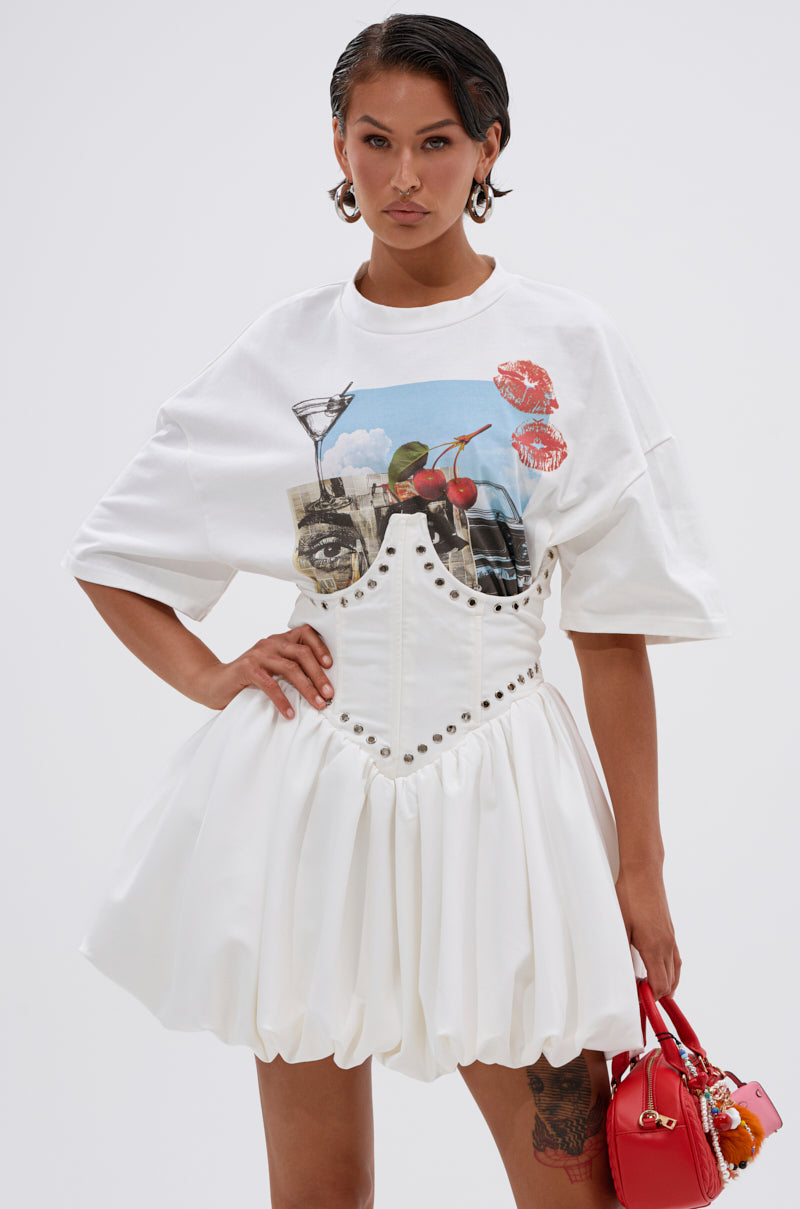 A woman poses in the COOL GIRL LOADING MINI DRESS WITH GRAPHIC TEE ATTACHED, featuring a corset-style bodice and oversized tee details. She completes the look with hoop earrings, a small red handbag, and slicked-back hair.