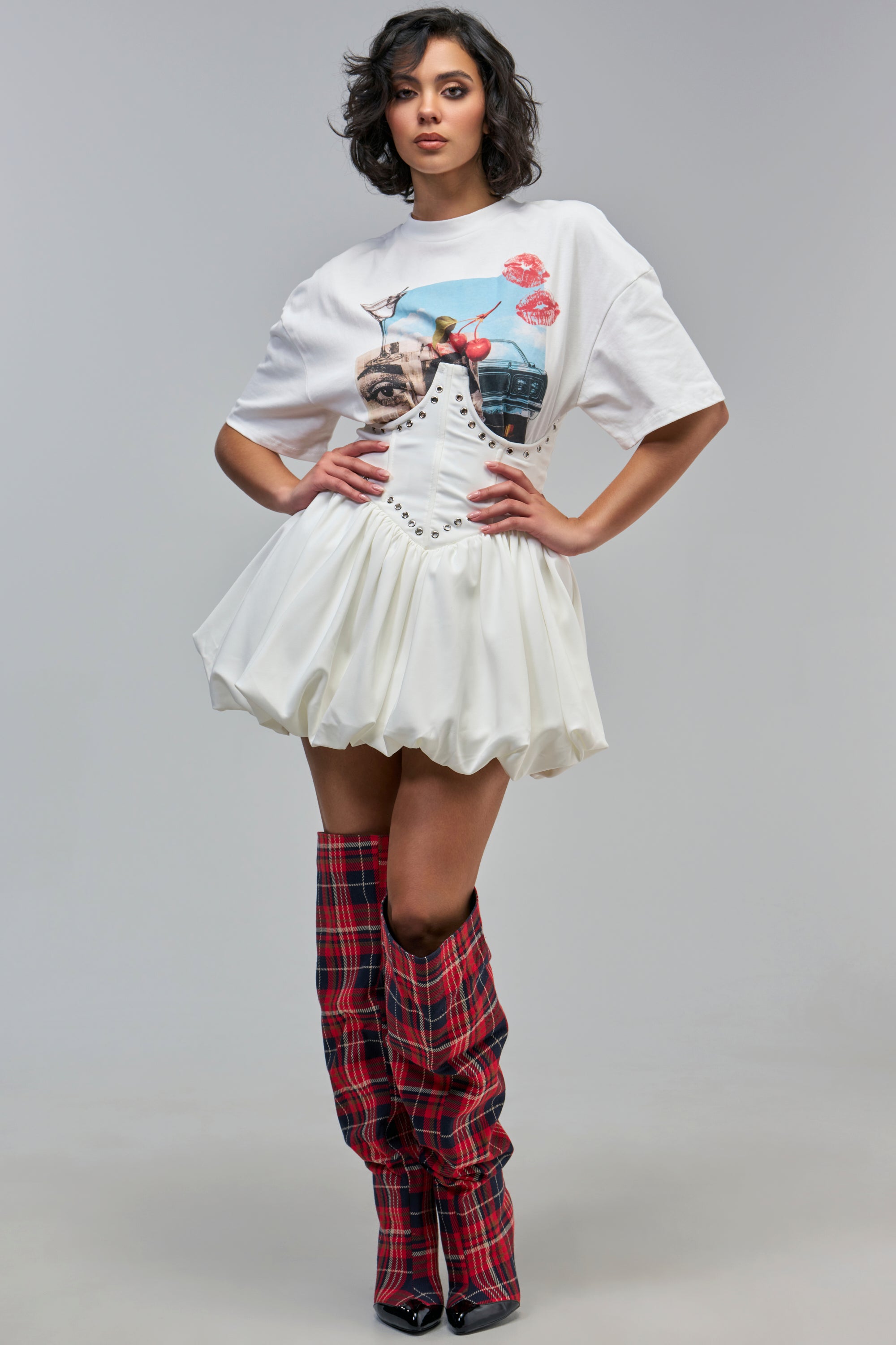 A person wearing a COOL GIRL LOADING MINI DRESS WITH GRAPHIC TEE ATTACHED, featuring a white corset bodice and red plaid over-the-knee boots.