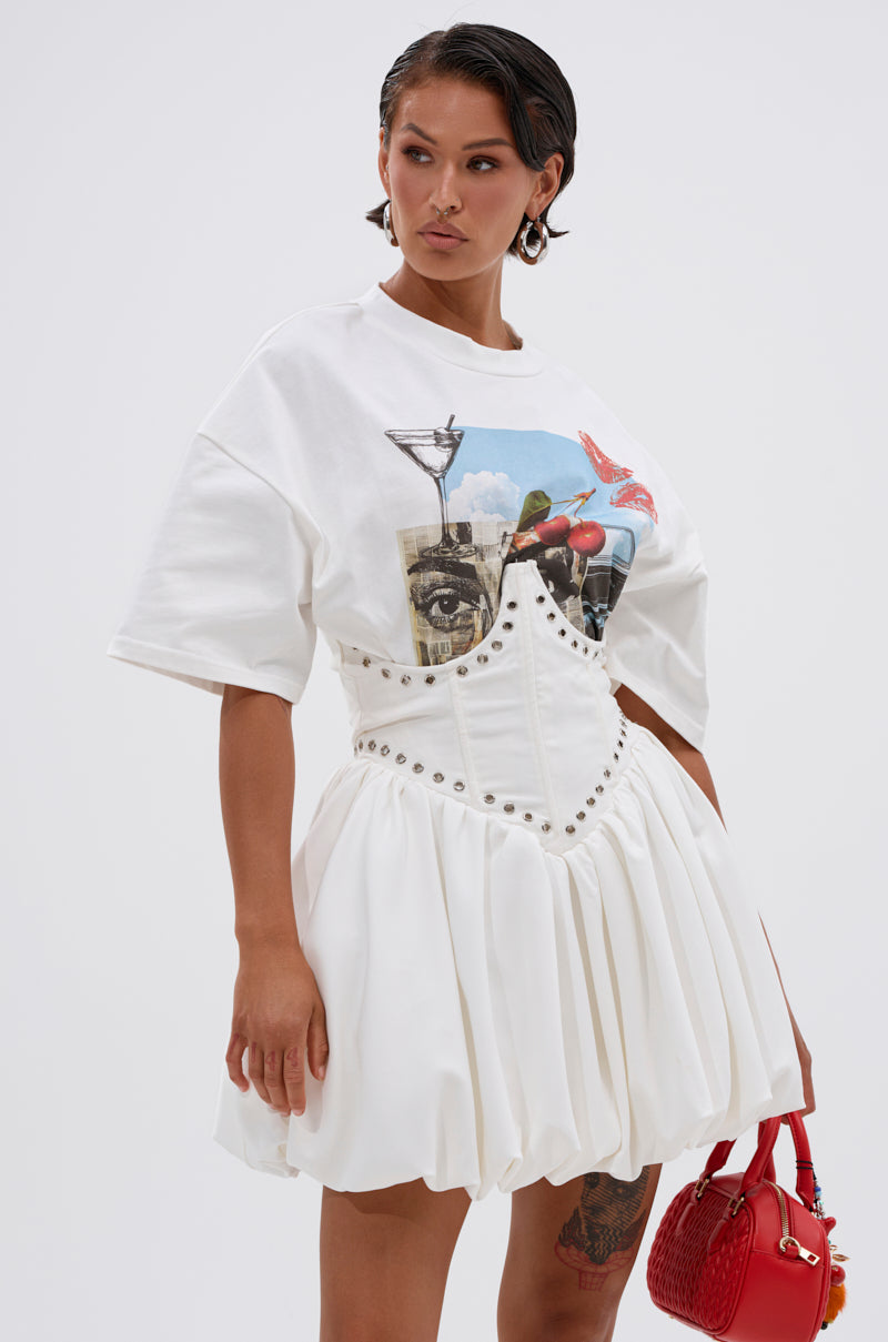 A woman with short dark hair and hoop earrings wears the COOL GIRL LOADING MINI DRESS WITH GRAPHIC TEE ATTACHED, featuring a corset-style bodice and flared skirt, and holds a small red handbag.