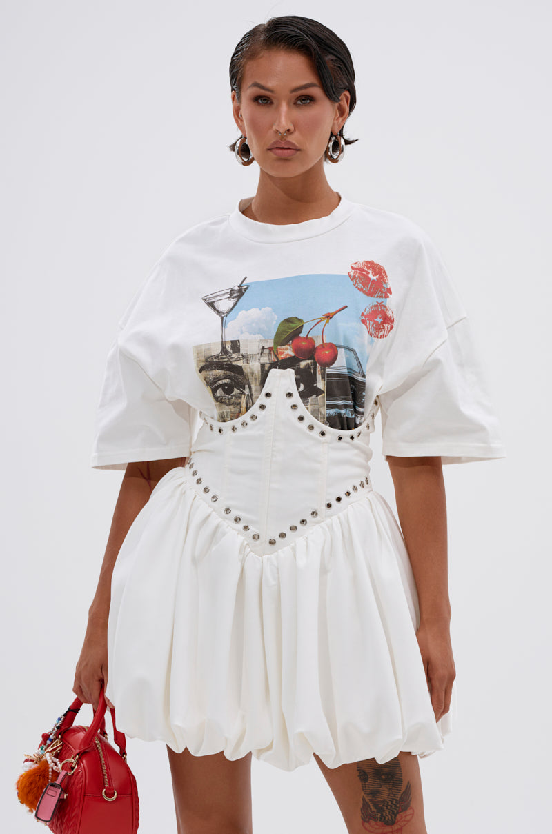A woman with short dark hair wears the COOL GIRL LOADING MINI DRESS WITH GRAPHIC TEE ATTACHED, featuring a cocktail and cherry print, styled with a red handbag and large hoop earrings.