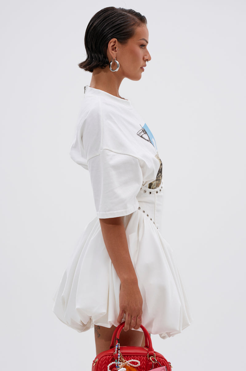 A woman with slicked-back dark hair wears the COOL GIRL LOADING MINI DRESS WITH GRAPHIC TEE ATTACHED and holds a red quilted handbag, posing against a plain white background.