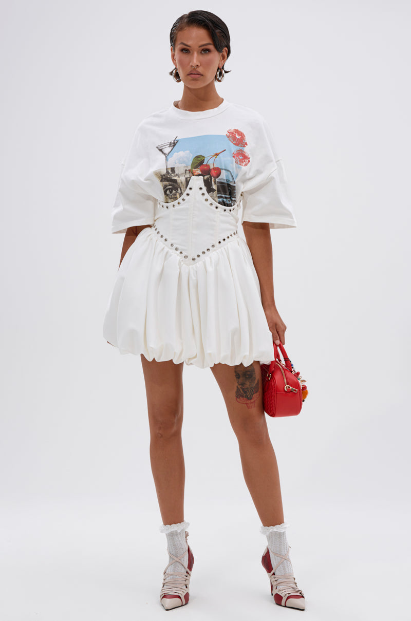 A woman with short, slicked-back hair stands against a plain background wearing the COOL GIRL LOADING MINI DRESS WITH GRAPHIC TEE ATTACHED, paired with white socks, open-toe heels, and a small red handbag.