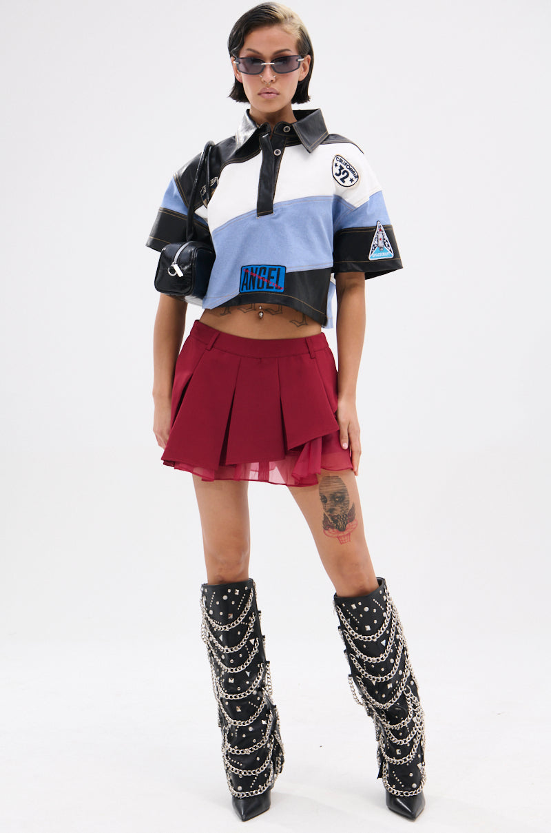 A person in sunglasses, a cropped blue and white collared shirt with patches, the AUDREY MINI PLEATED SKIRT in red, and tall black boots with silver chains. They have a large thigh tattoo and stand against a plain white background. 5