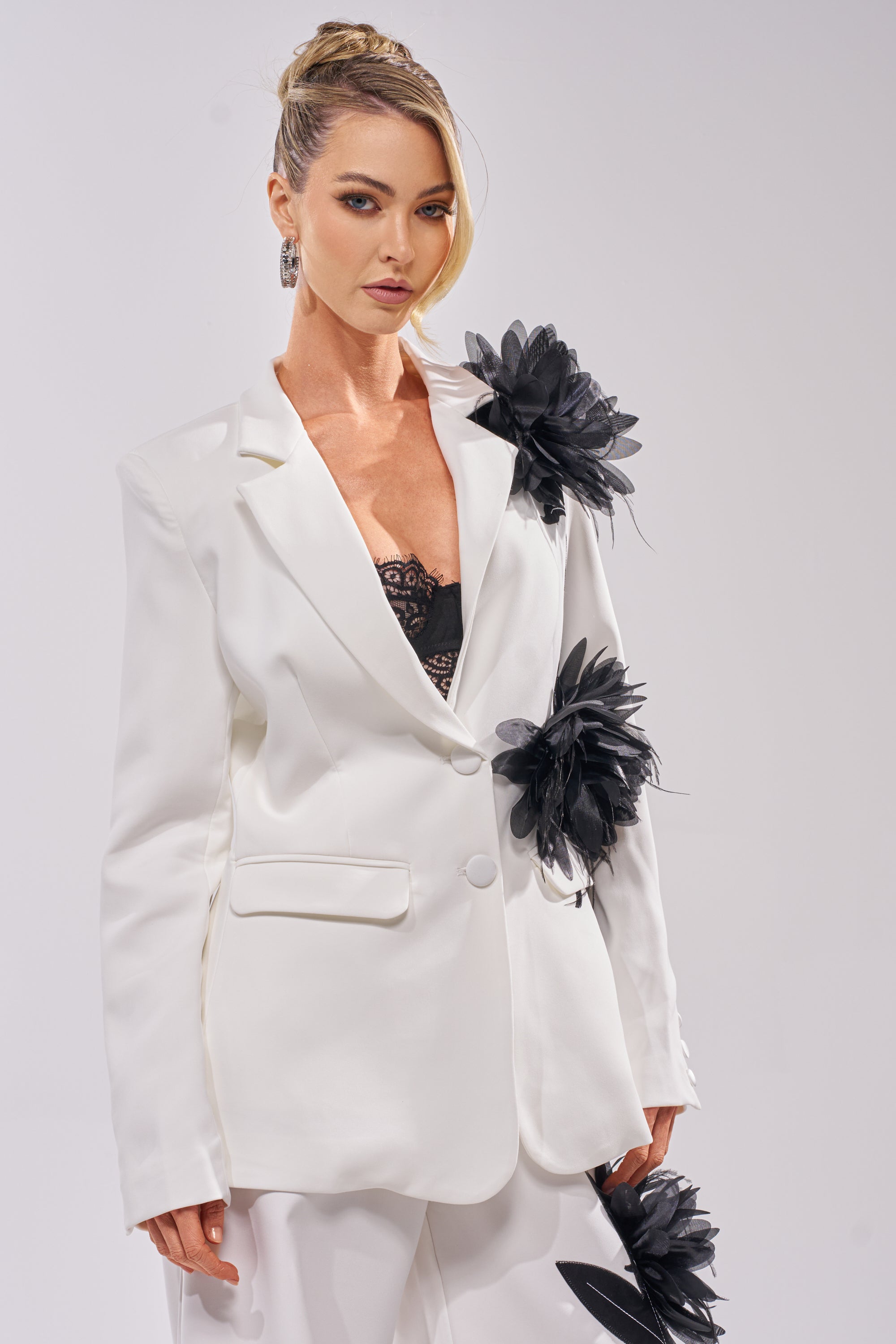 A woman wears the IN FULL BLOOM APPLIQUE BLAZER over a black lace top with matching white pants. Her hair is styled in an elegant updo as she stands against a plain light background. 0