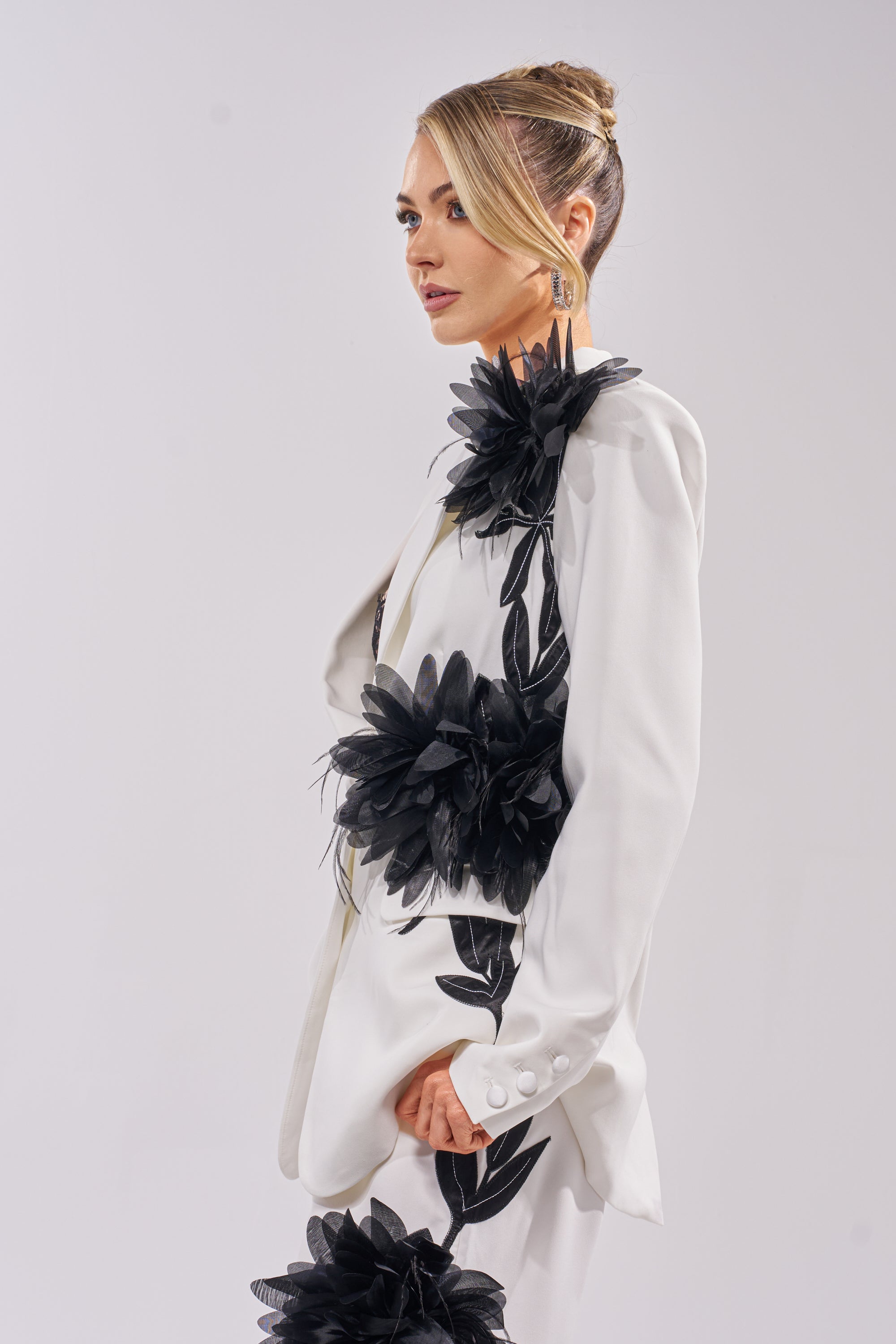 A woman with blonde hair in a sleek bun models the IN FULL BLOOM APPLIQUE BLAZER, a white women's suiting blazer decorated with bold black floral and leaf appliqués, posing in profile against a plain light background. 3