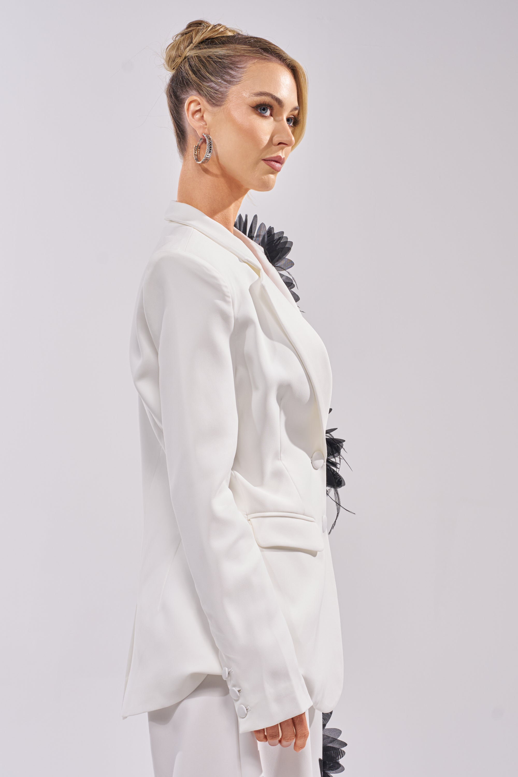 A blonde woman with a sleek updo wears the IN FULL BLOOM APPLIQUE BLAZER, an elegant white piece with black floral 3D details, shown in profile against a plain light background. 4