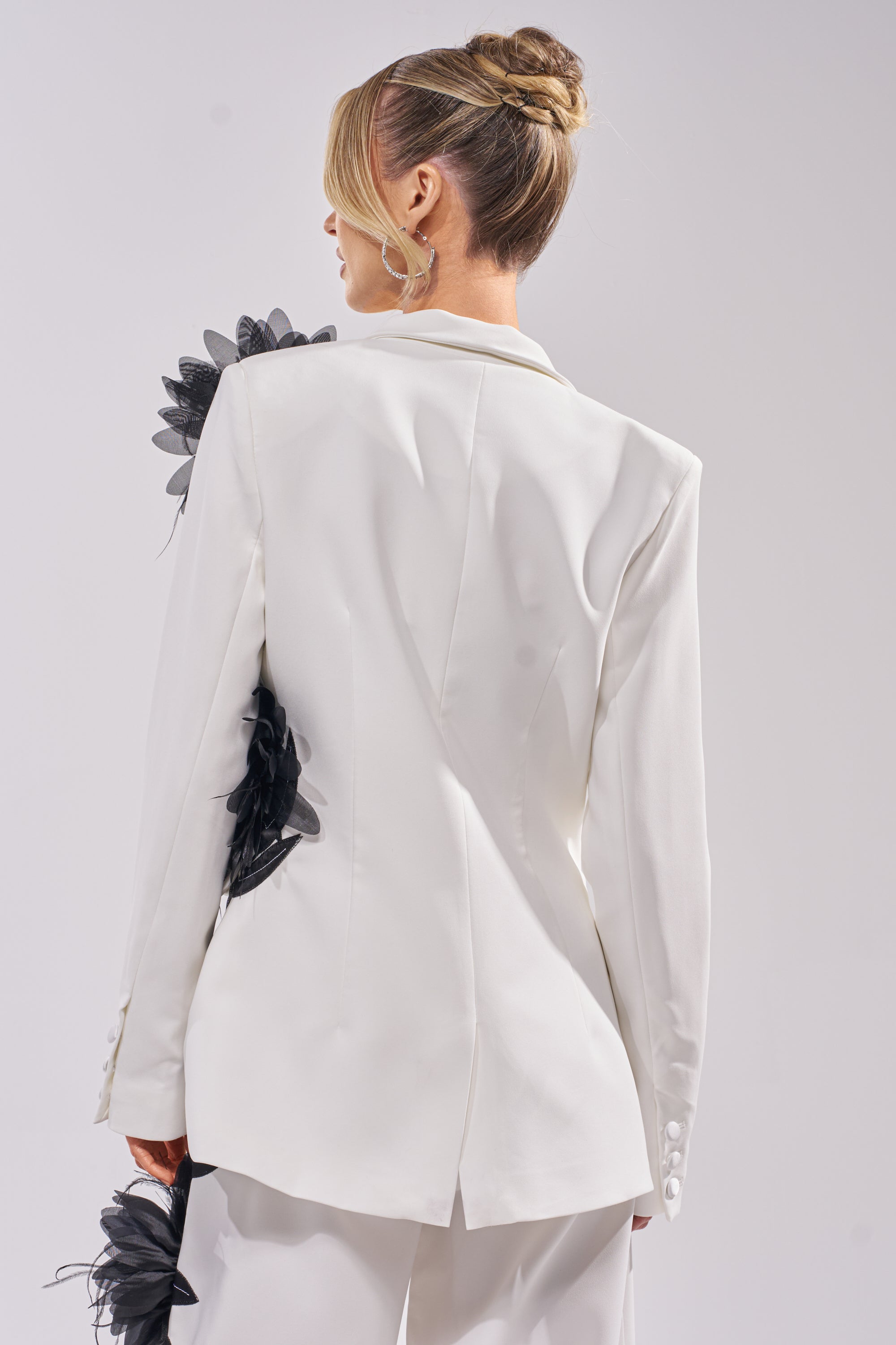A woman with blonde hair in a neat bun, seen from behind, wears the IN FULL BLOOM APPLIQUE BLAZER and matching pants. Black 3D floral appliques decorate her left shoulder and lower back against a plain light background. 5