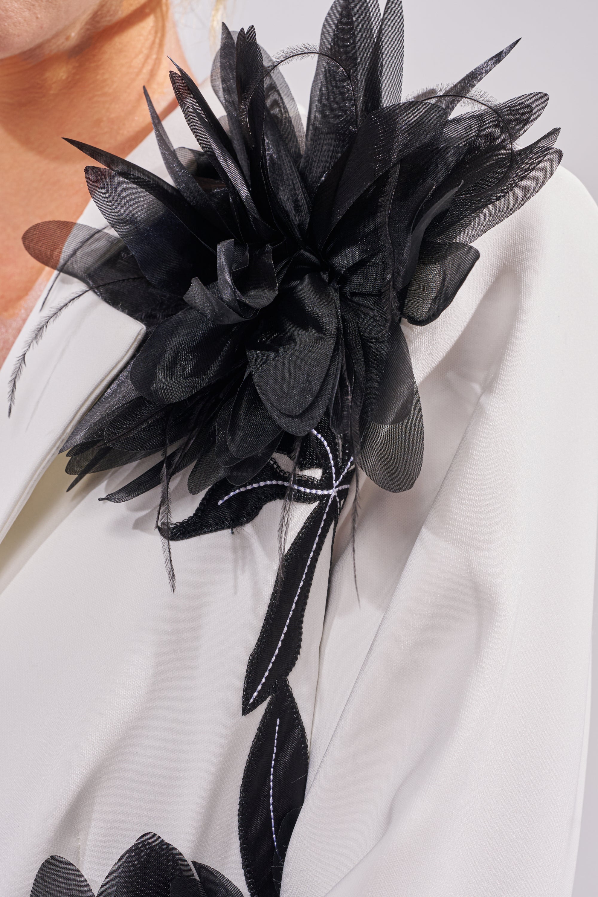 A close-up of a large black fabric flower brooch with layered petals and ribbons, pinned to the lapel of the IN FULL BLOOM APPLIQUE BLAZER worn by a person. 8
