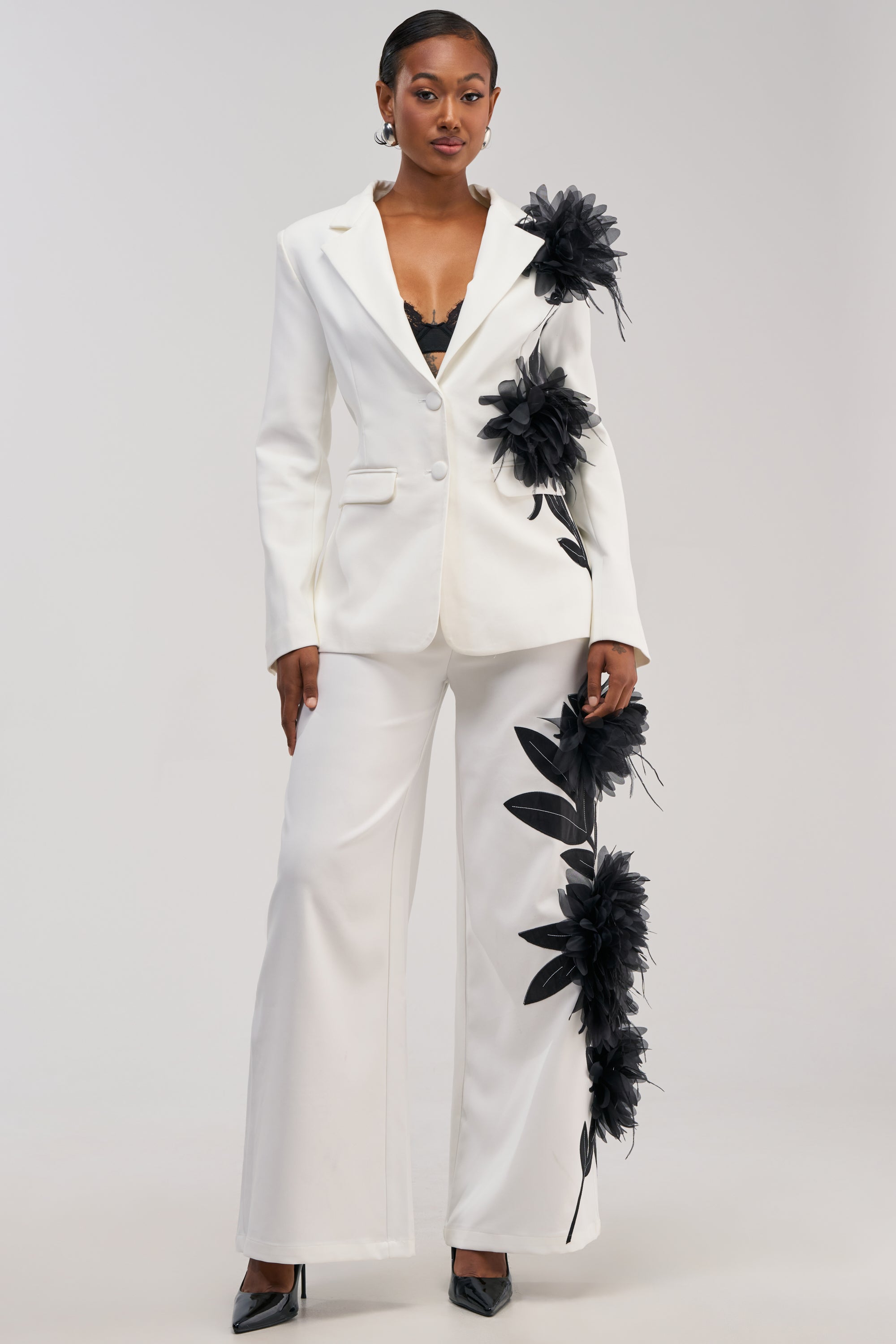 A woman models the IN FULL BLOOM APPLIQUE TROUSER, paired with a matching white jacket adorned with large black floral embroidery and leaf appliqués, a visible black lace top, and black heels. She poses against a plain light background.