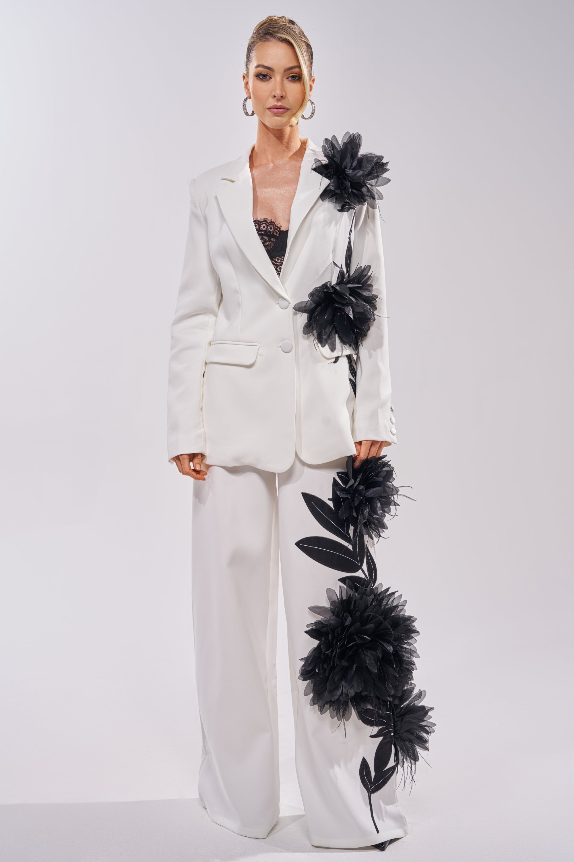 A woman wears the IN FULL BLOOM APPLIQUE TROUSER, a white wide-leg suit with oversized black floral embroidery on the jacket and trousers. She pairs it with a black lace top and stands confidently against a plain background. 0