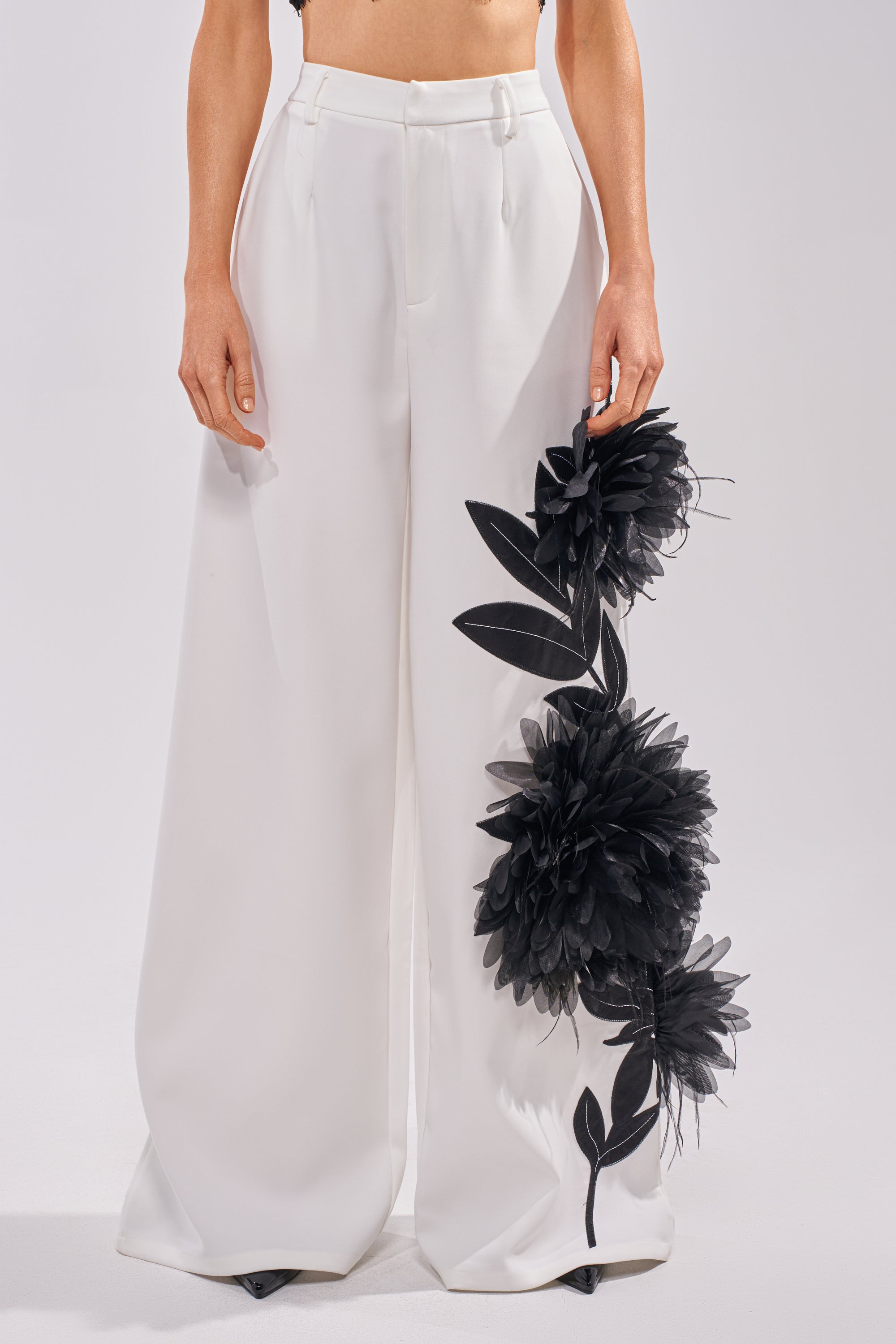 Model wears the IN FULL BLOOM APPLIQUE TROUSER—high-rise wide-leg pants featuring large black floral appliqués on one leg—standing against a plain white background with the torso cropped out. 1