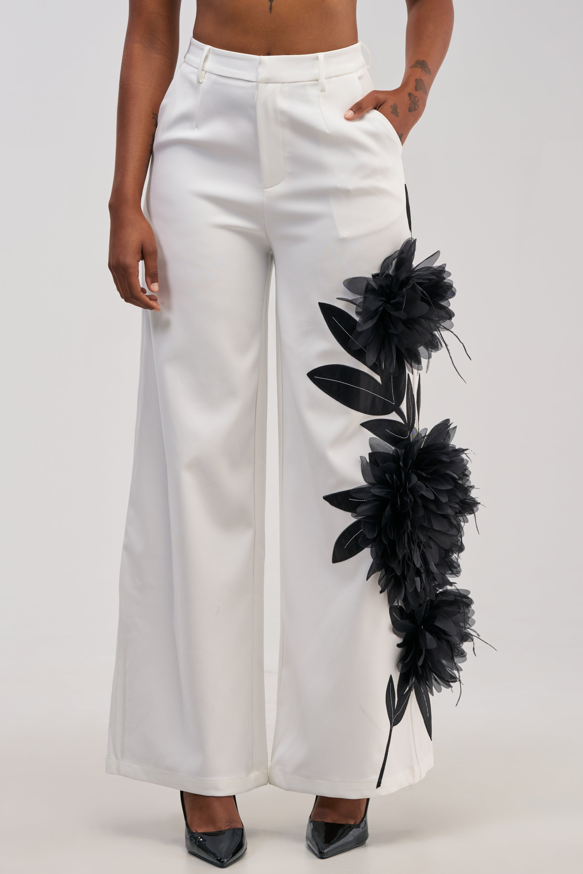 A person wears the IN FULL BLOOM APPLIQUE TROUSER—high-waisted white trousers featuring striking large black tulle floral appliqués on one leg—paired elegantly with black pointed-toe heels. The photo is cropped from mid-torso to feet and set against a plain background.