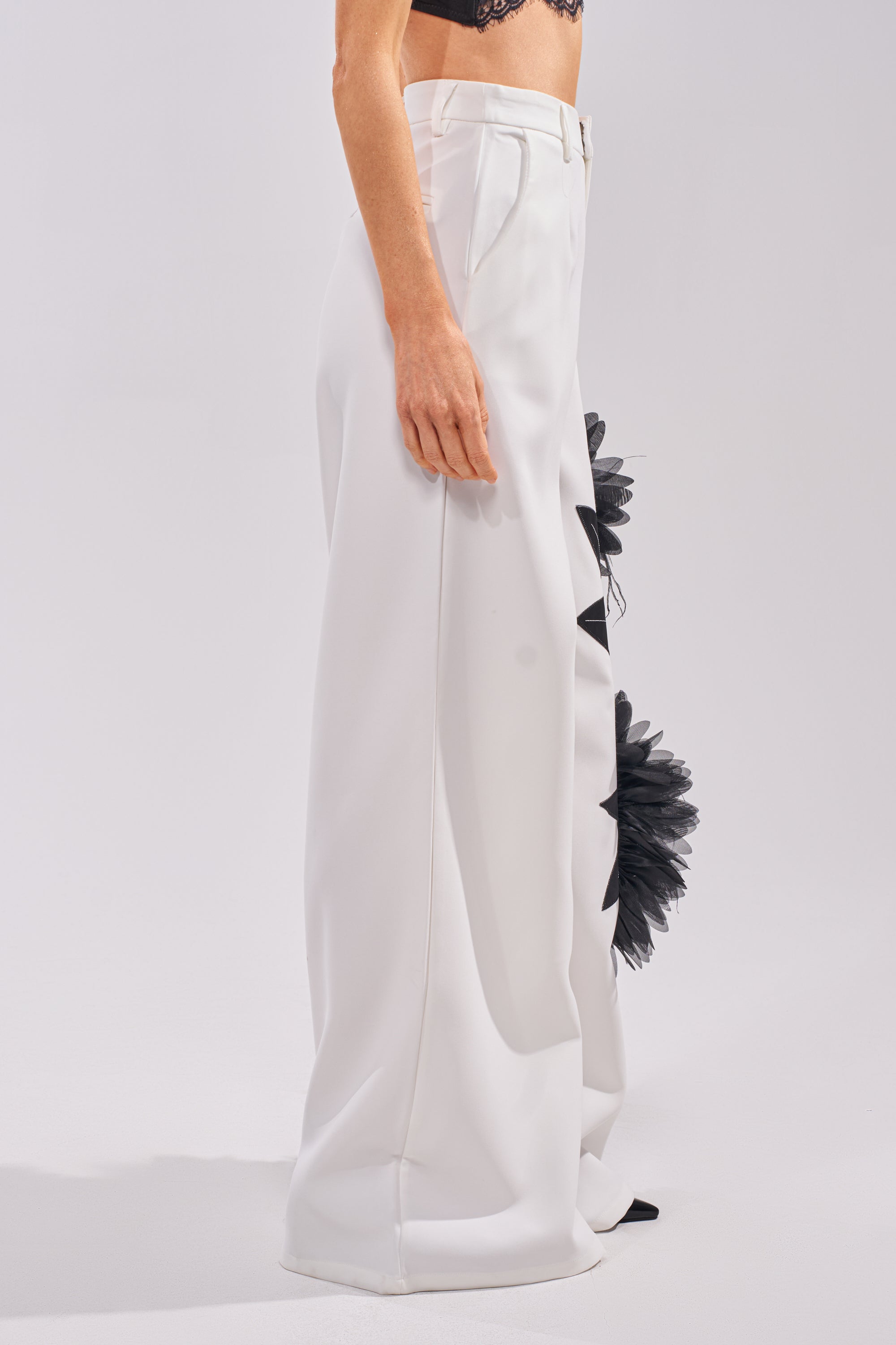 A person wears the IN FULL BLOOM APPLIQUE TROUSER—high-waisted, wide-leg with black floral tulle accents—and a black lace bralette, standing against a plain white background. 2