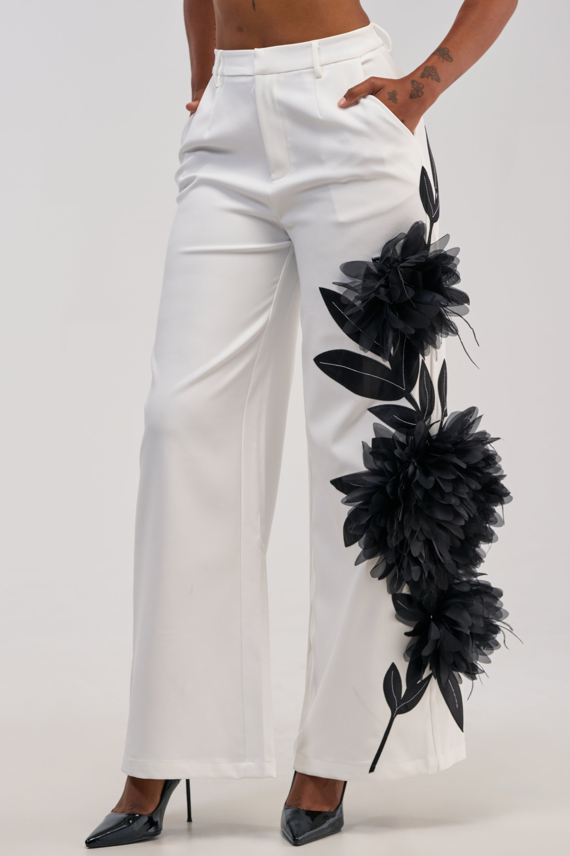 A woman wears the IN FULL BLOOM APPLIQUE TROUSER—white high-waisted wide-leg pants featuring a bold black tulle flower appliqué on one leg. She stands with her hands in her pockets, paired with black high heels and a visible forearm tattoo.