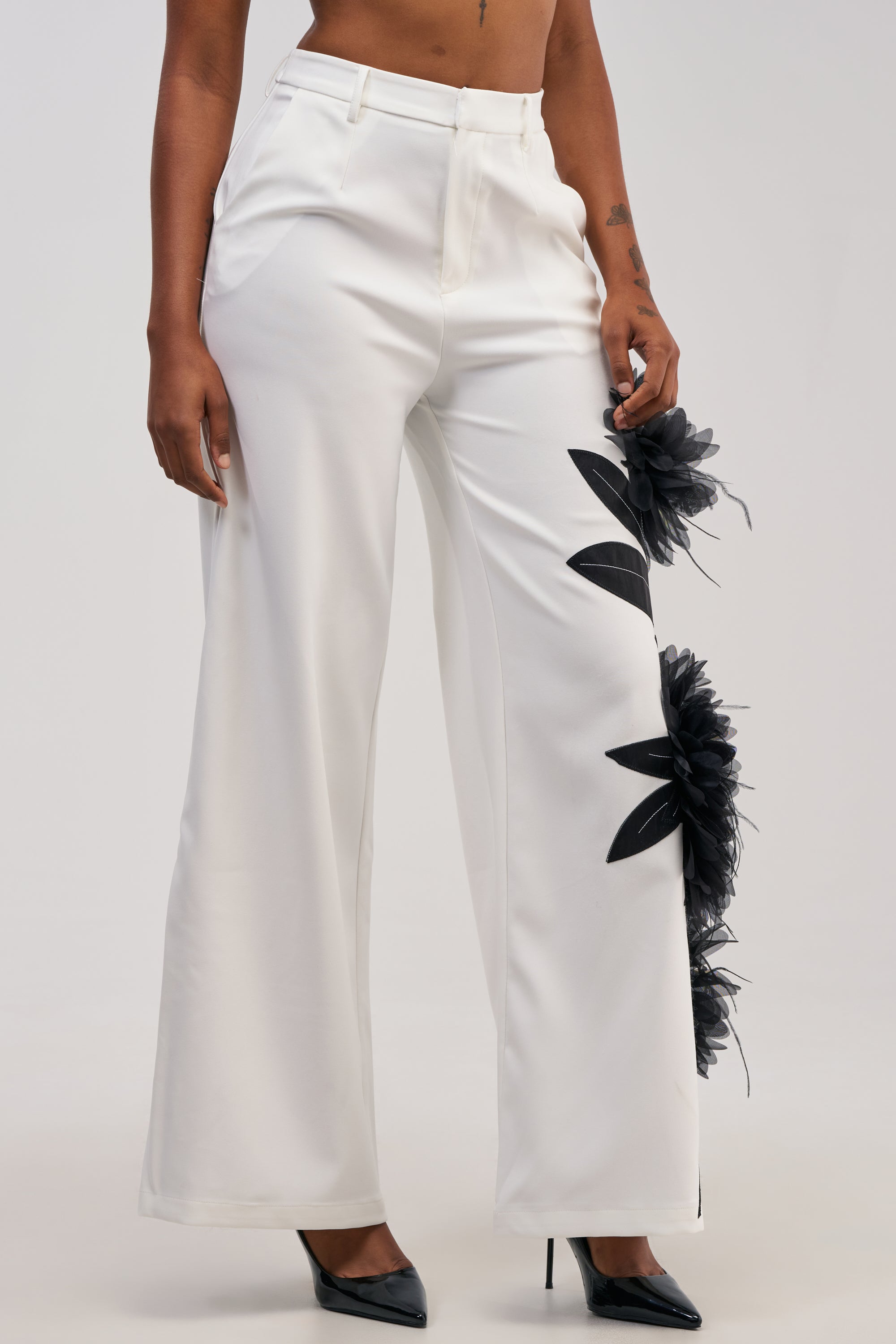 A person wearing the IN FULL BLOOM APPLIQUE TROUSER, featuring high-waisted white trousers with intricate black tulle flower appliqués on one leg, paired with black pointed-toe heels.