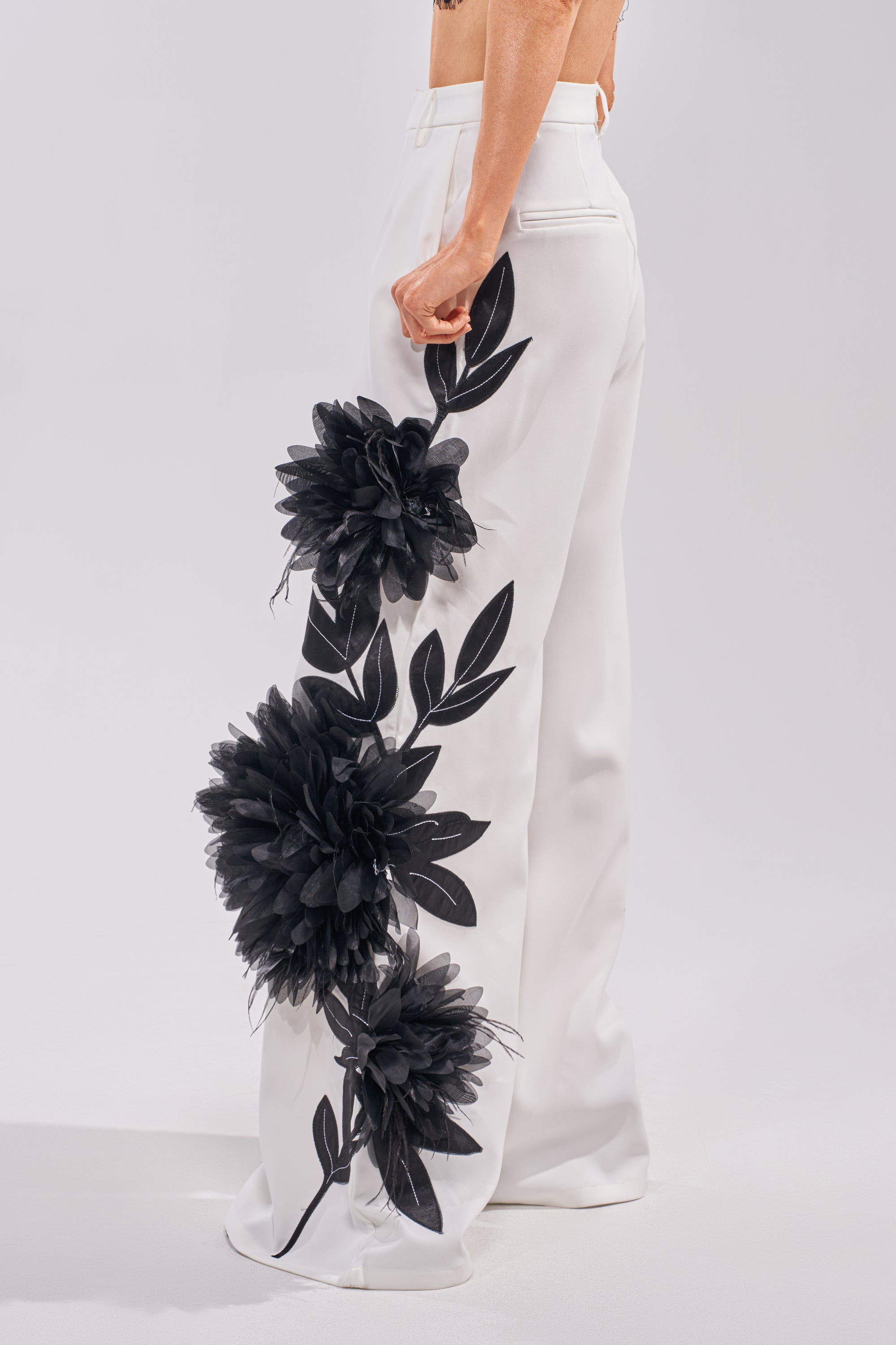 A model showcases the IN FULL BLOOM APPLIQUE TROUSER—high-waisted white trousers with bold black floral and leaf embroidery along the sides, featuring a stylish wide-leg silhouette, seen from the waist down against a plain background. 3