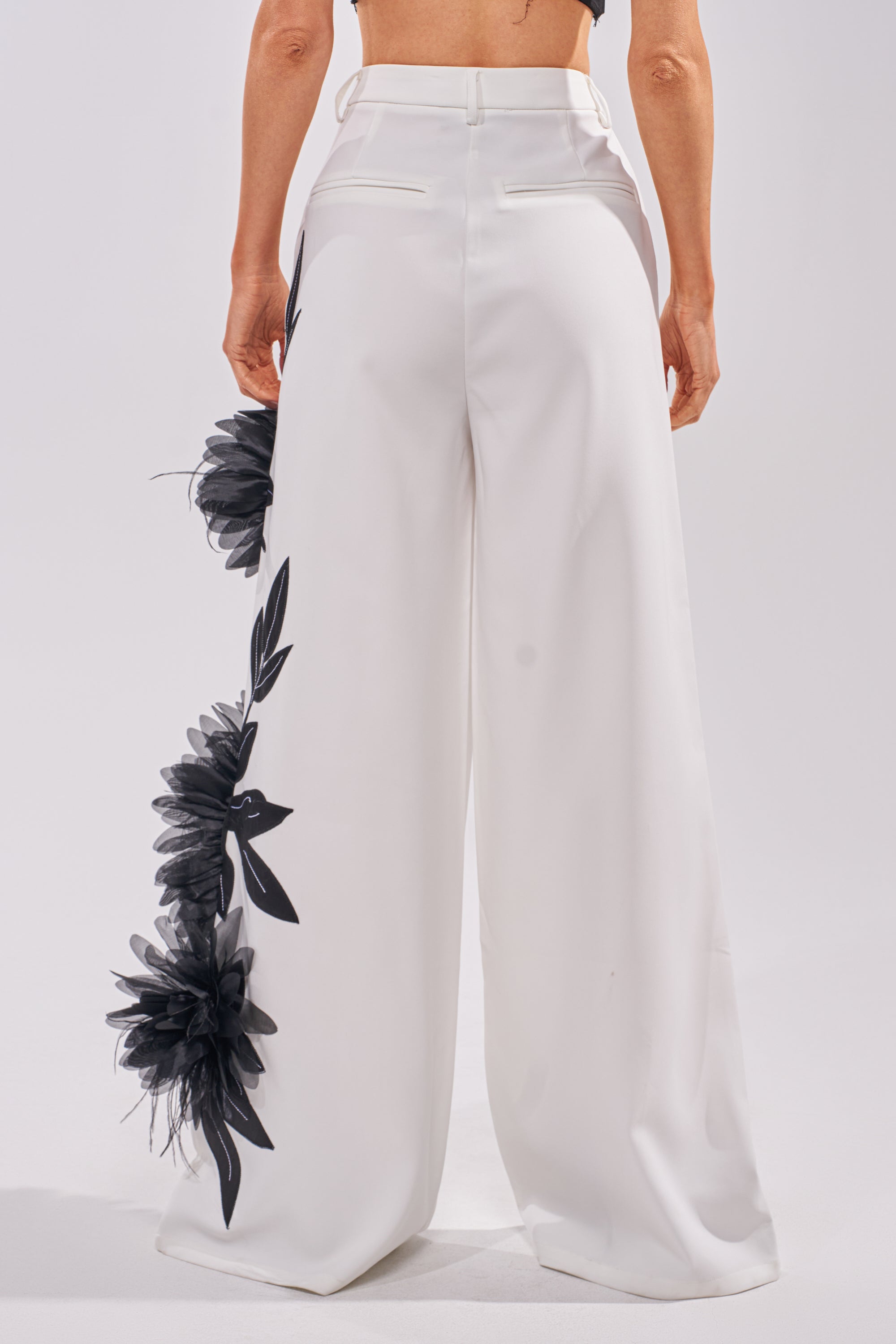Wearing the IN FULL BLOOM APPLIQUE TROUSER, high-waisted white pants with wide legs and black tulle flower appliqué on one leg, styled from the back with a black cropped top. 4