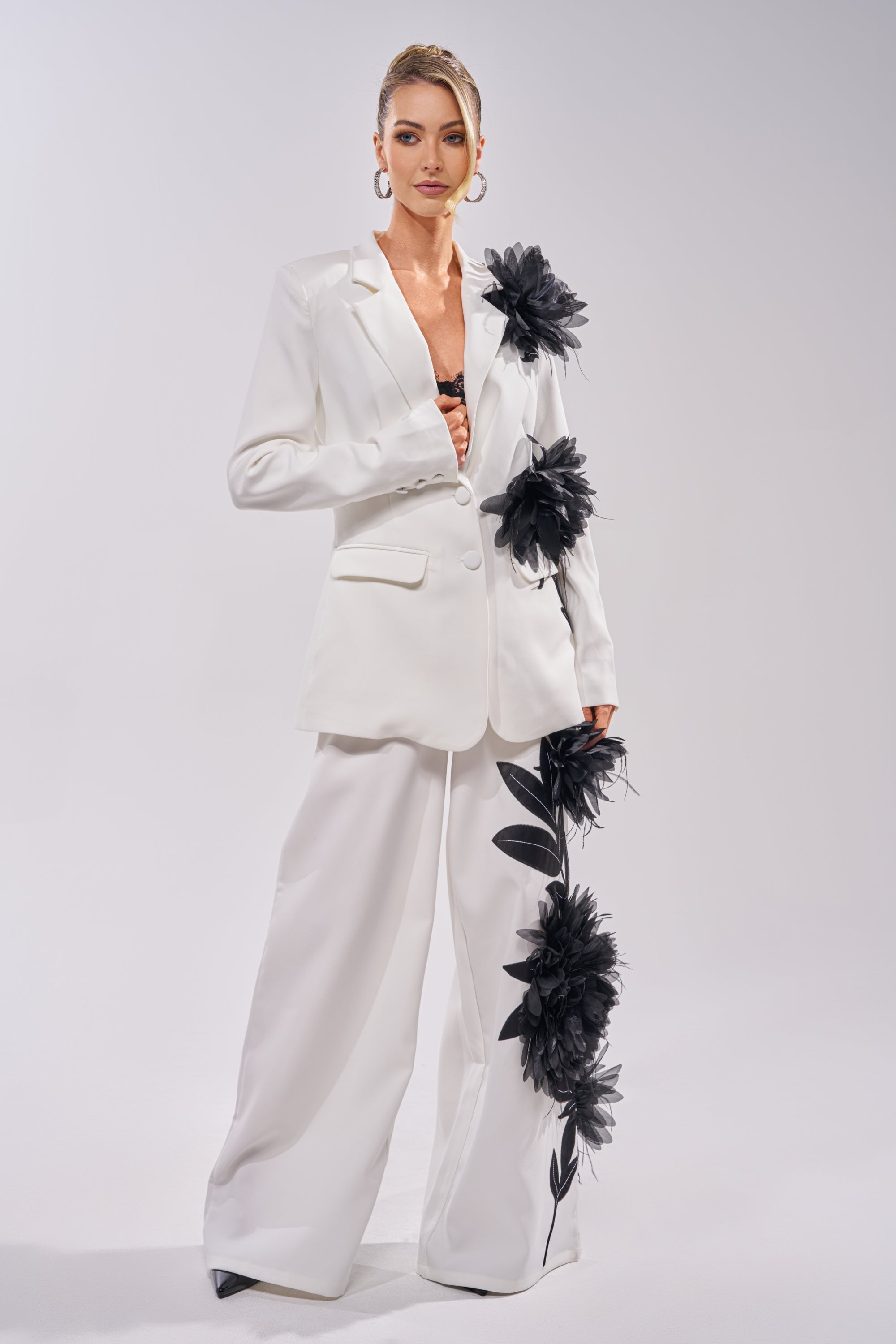 A woman wears the IN FULL BLOOM APPLIQUE TROUSER—a white pantsuit with wide-leg trousers and bold black floral appliqué—paired with hoop earrings and black heels, standing against a plain light background. 5