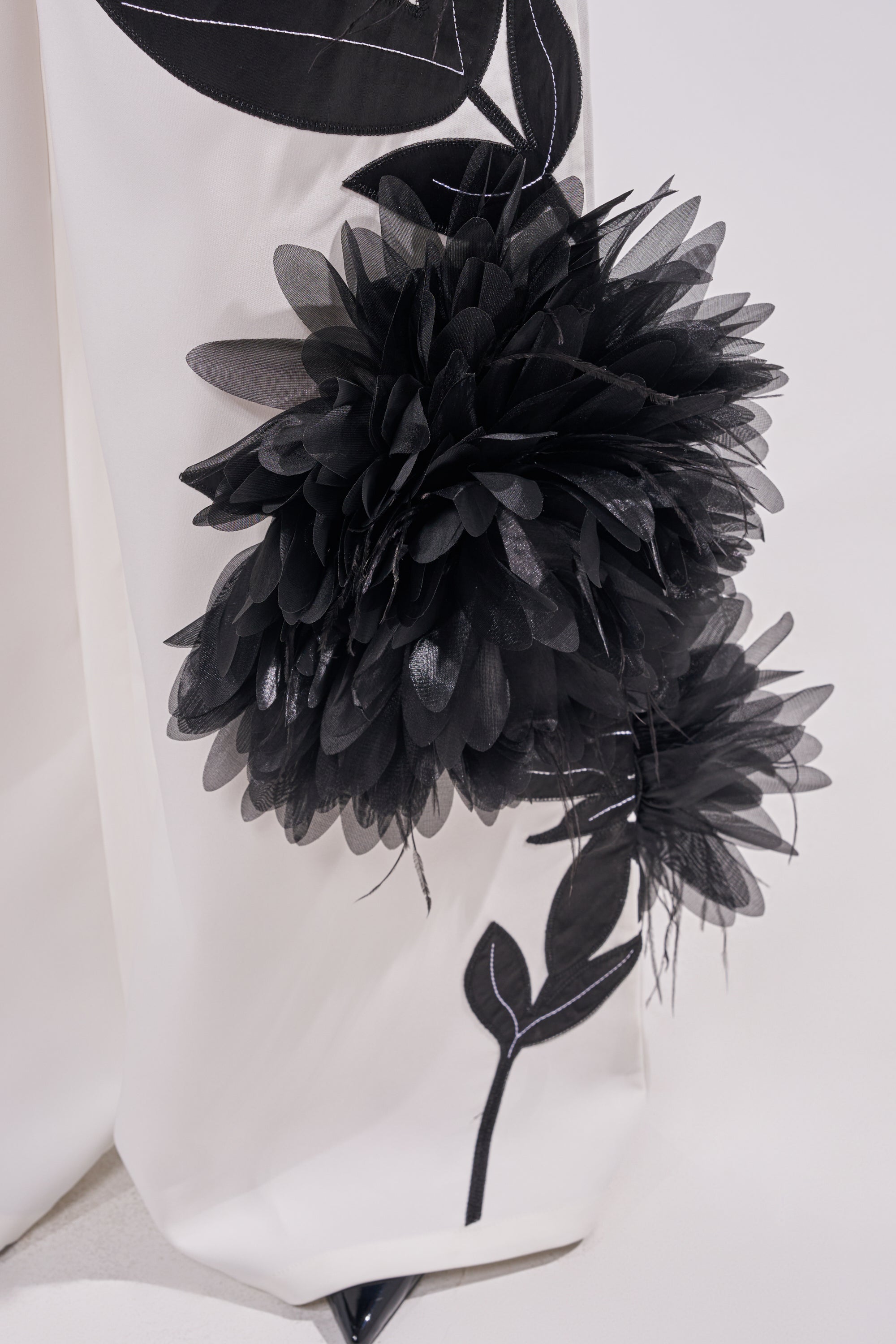 Close-up of the IN FULL BLOOM APPLIQUE TROUSER: white wide-leg trousers decorated with bold, textured black floral embroidery and leaf appliqué. Layered petals add dimension; a pointed black shoe peeks out at the hem. 6