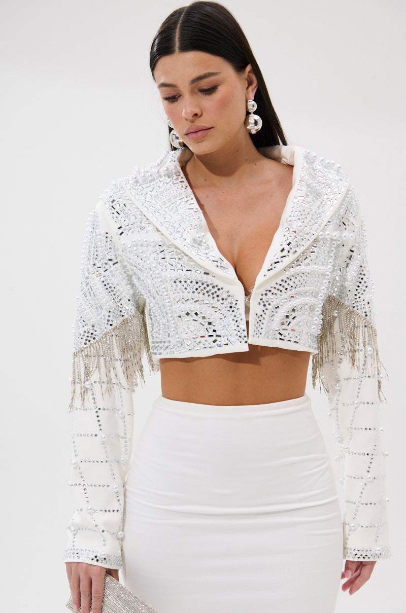 A woman with long dark hair models the DIRTY MARTINI EMBELLISHED BLAZER IN WHITE, featuring sparkling pearls and rhinestones with silver fringe, over a fitted white skirt. She looks down, showcasing statement earrings against a plain light background.