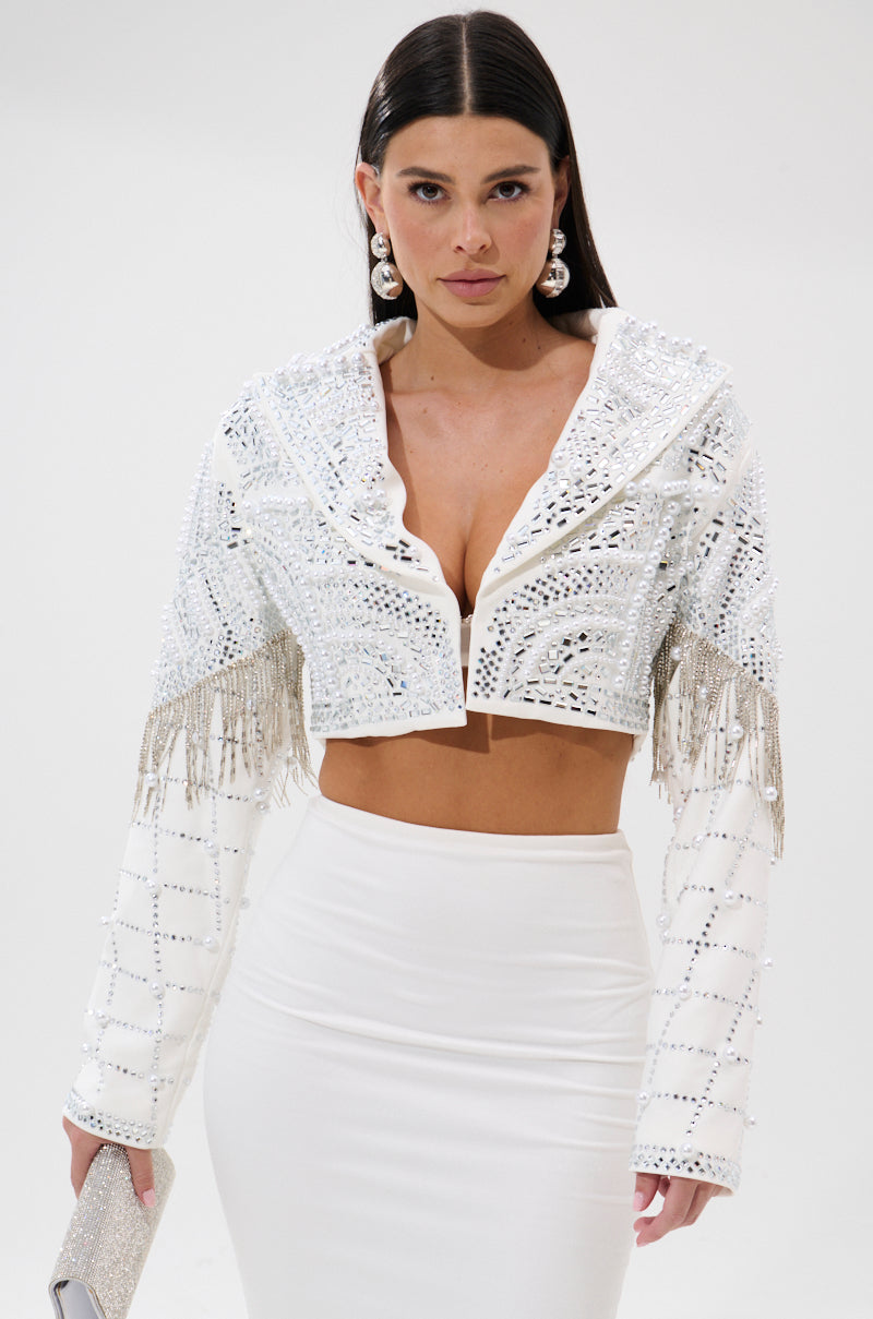 A woman with long, straight hair and statement earrings stands against a plain background, holding a glittery clutch. She wears a fitted high-waisted skirt and the DIRTY MARTINI EMBELLISHED BLAZER IN WHITE with fringe details.