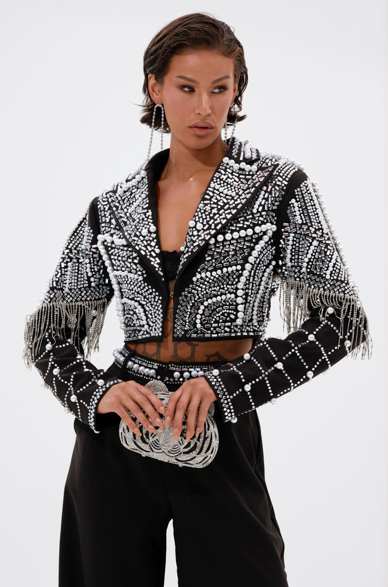 A person wears the DIRTY MARTINI EMBELLISHED BLAZER, a cropped black blazer with silver beading and fringe, paired with high-waisted black pants and a matching beaded clutch against a plain background.