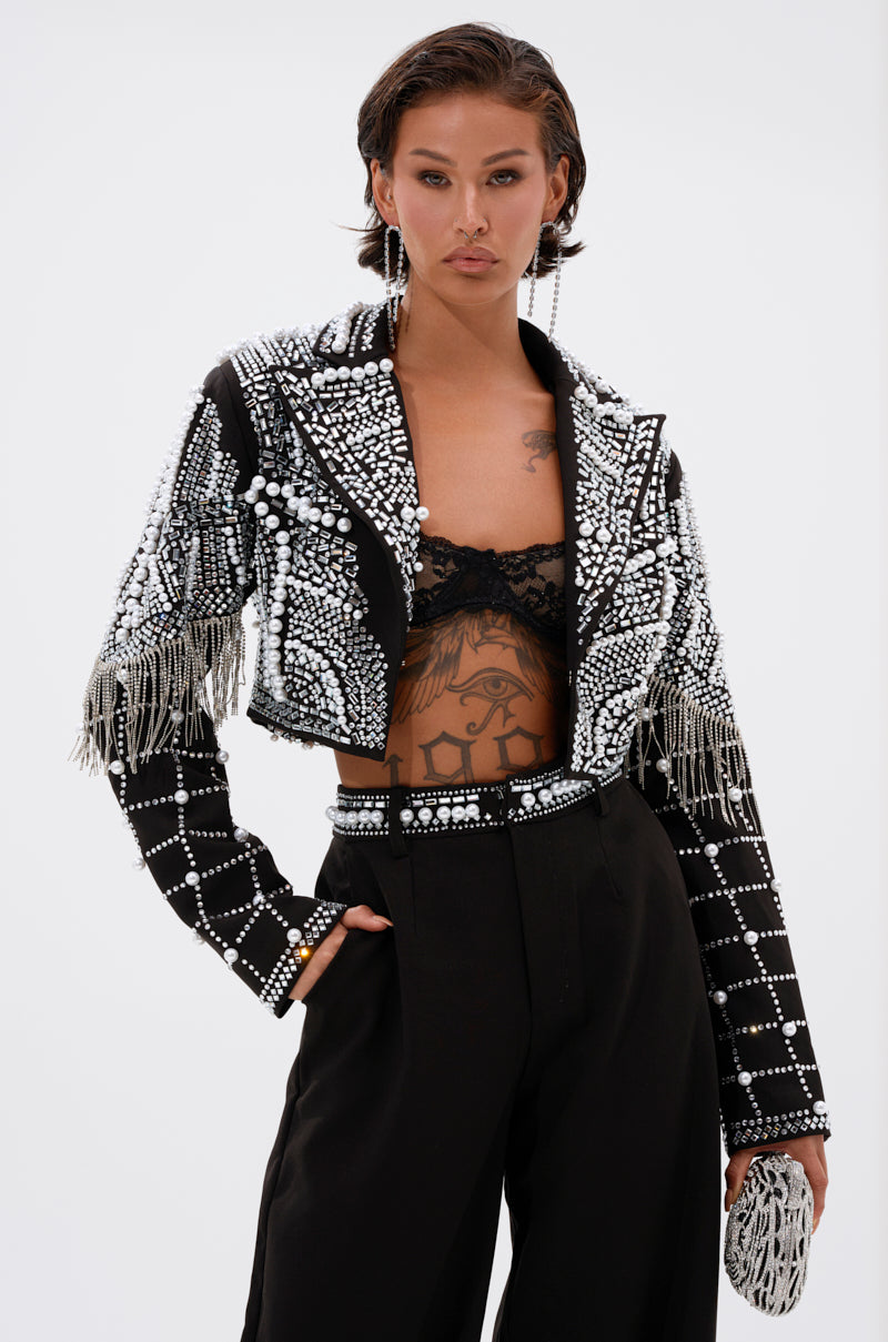 A person models the DIRTY MARTINI EMBELLISHED BLAZER—cropped, black, with pearl rhinestones, white beads, and fringe—styled with high-waisted black pants and a lace bralette against a plain white background.
