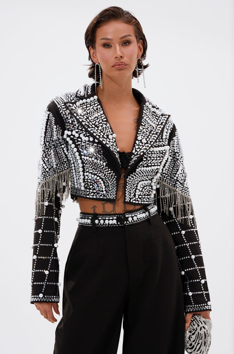 A person with short dark hair wears the DIRTY MARTINI EMBELLISHED BLAZER adorned with silver mirrors, sequins, and fringe, paired with black high-waisted pants, dangling earrings, and a matching clutch against a plain background.
