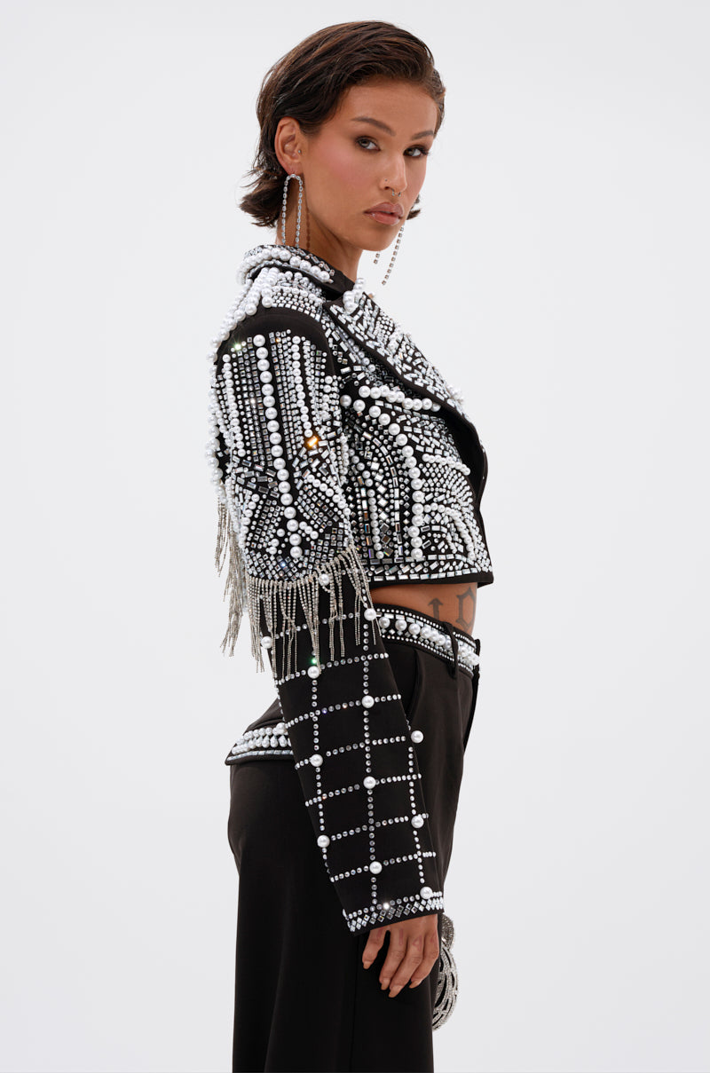 A woman with short styled brown hair faces sideways, wearing the DIRTY MARTINI EMBELLISHED BLAZER with black high-waisted pants and large hoop earrings. The blazer features black pearls, rhinestones, and silver fringes.