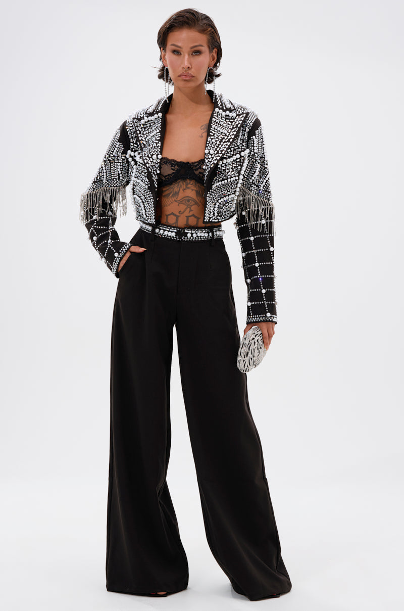 A person stands confidently in wide-leg black pants, a black lace bralette, and the DIRTY MARTINI EMBELLISHED BLAZER with pearl rhinestones and fringe. They hold a matching clutch and have short, slicked-back hair.