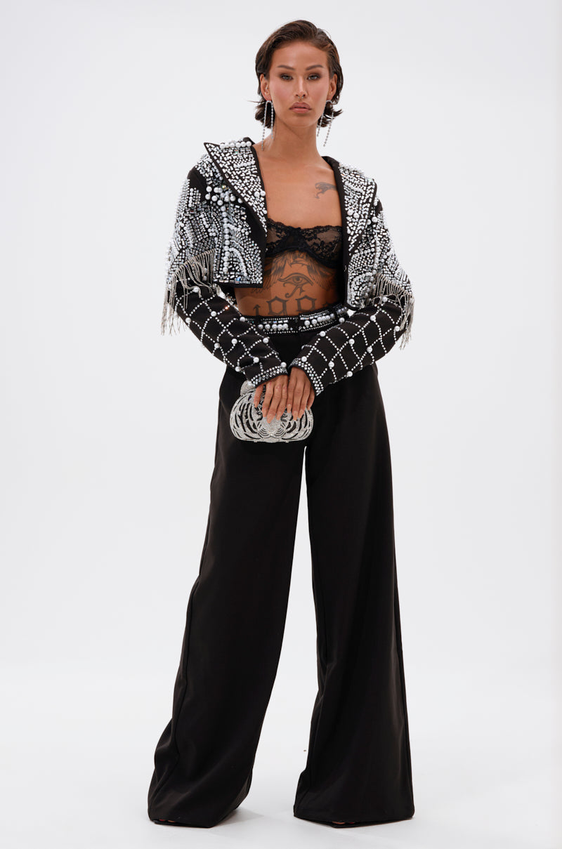 A person stands confidently in wide-leg black pants, a black lace bralette, and the DIRTY MARTINI EMBELLISHED BLAZER with silver accents and fringe. They hold a round silver clutch and display visible torso tattoos.