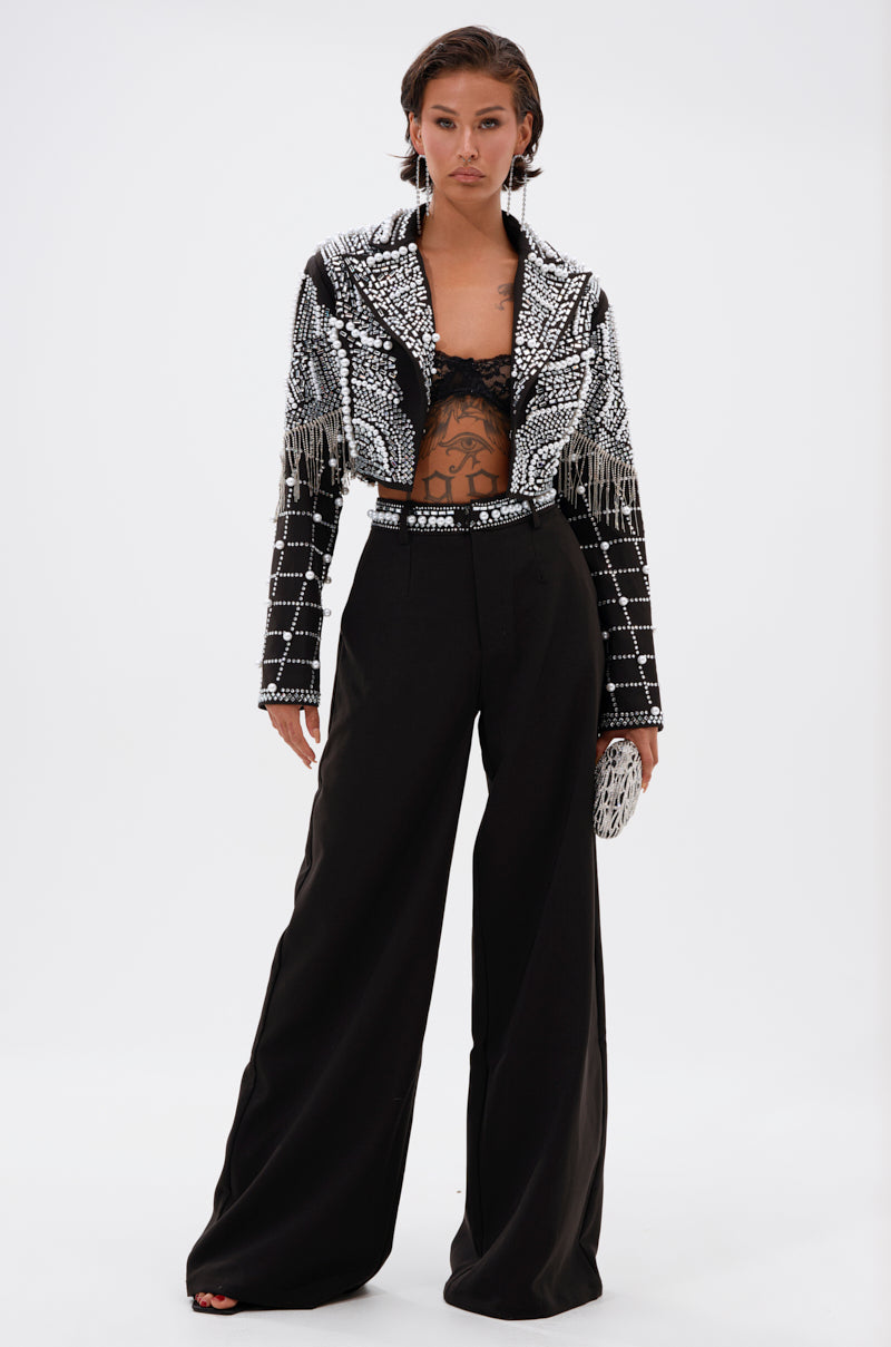 A woman stands confidently in wide-leg black pants, a lacy black bralette, and the DIRTY MARTINI EMBELLISHED BLAZER with pearl rhinestone fringe. She holds a small clutch and wears statement earrings, her hair styled short and slicked-back.