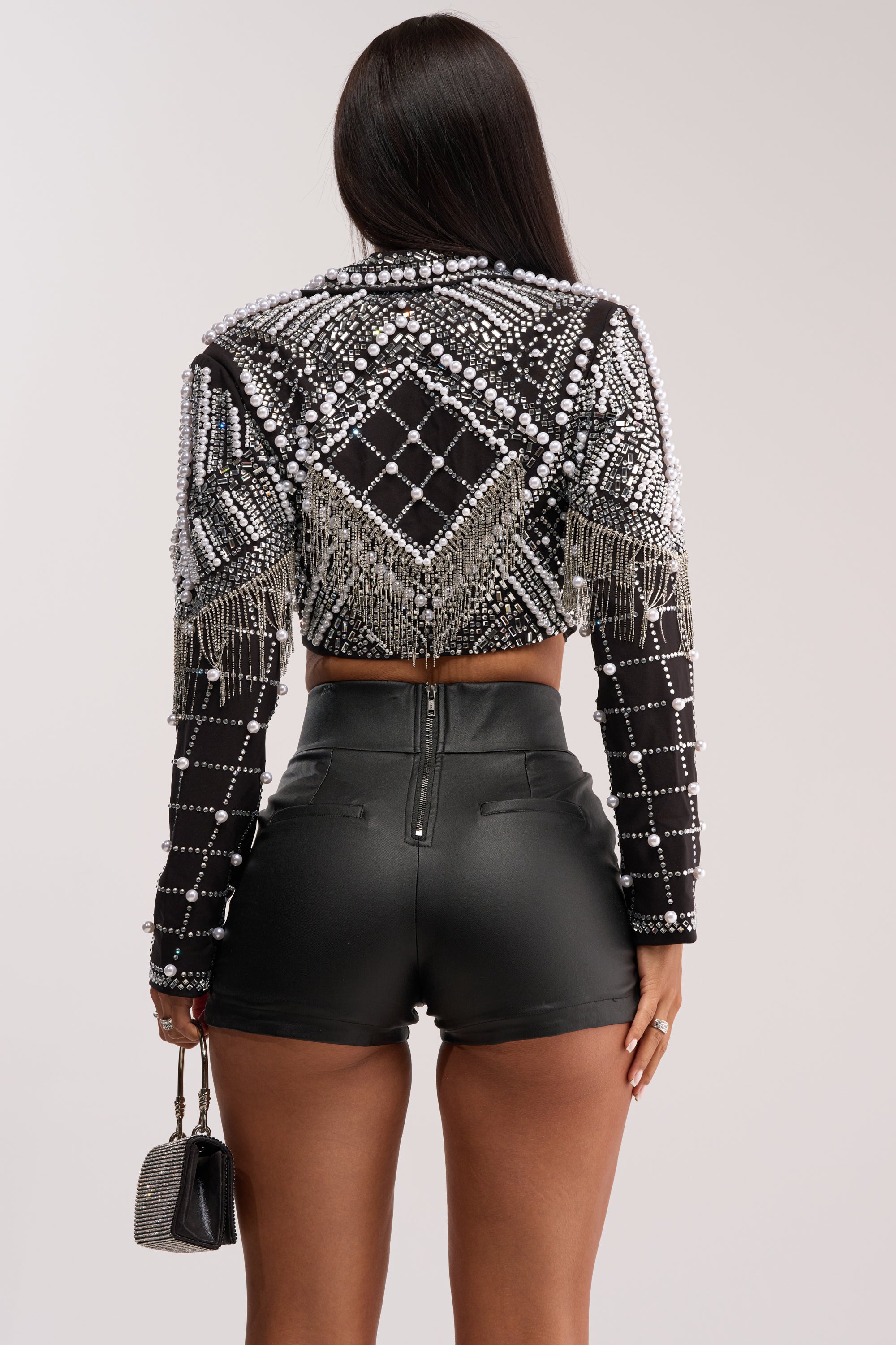 A woman faces away, wearing the DIRTY MARTINI EMBELLISHED BLAZER with fringe and high-waisted black leather shorts. She holds a glittery handbag in her left hand, her long straight hair loose down her back.