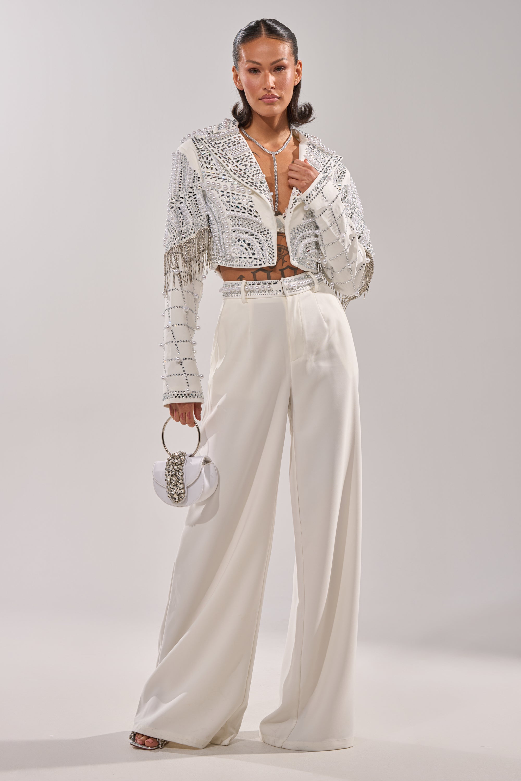 A woman models the DIRTY MARTINI EMBELLISHED TROUSER IN WHITE—high-rise wide-leg pants paired with a studded, fringed cropped jacket and crop top. She accessorizes with a small white ring-handle handbag and hoop earrings. 0