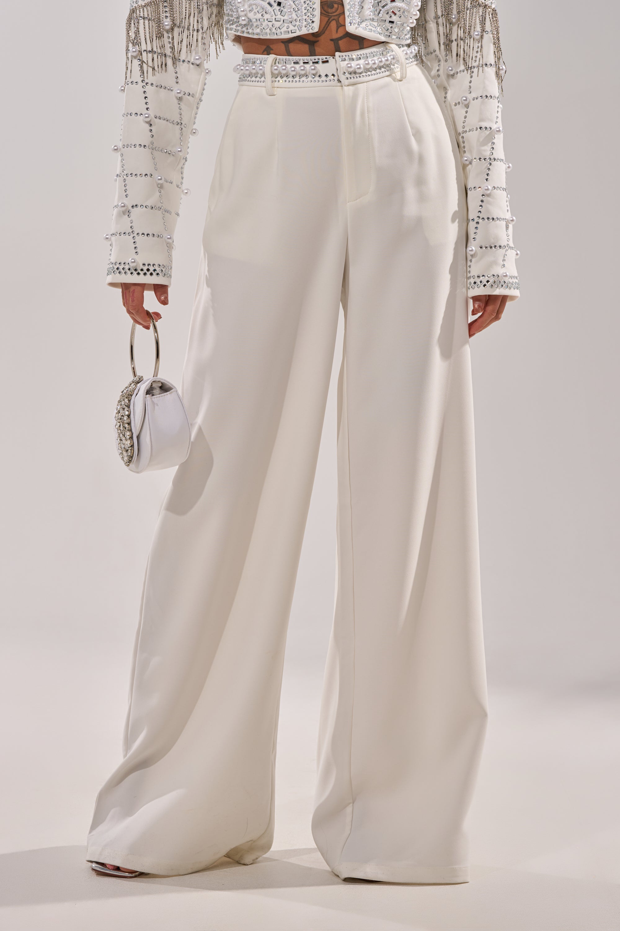 Wearing DIRTY MARTINI EMBELLISHED TROUSER IN WHITE, a high-waisted, wide-leg style, with a beaded long-sleeve top and white purse, this person stands against a plain background. 1