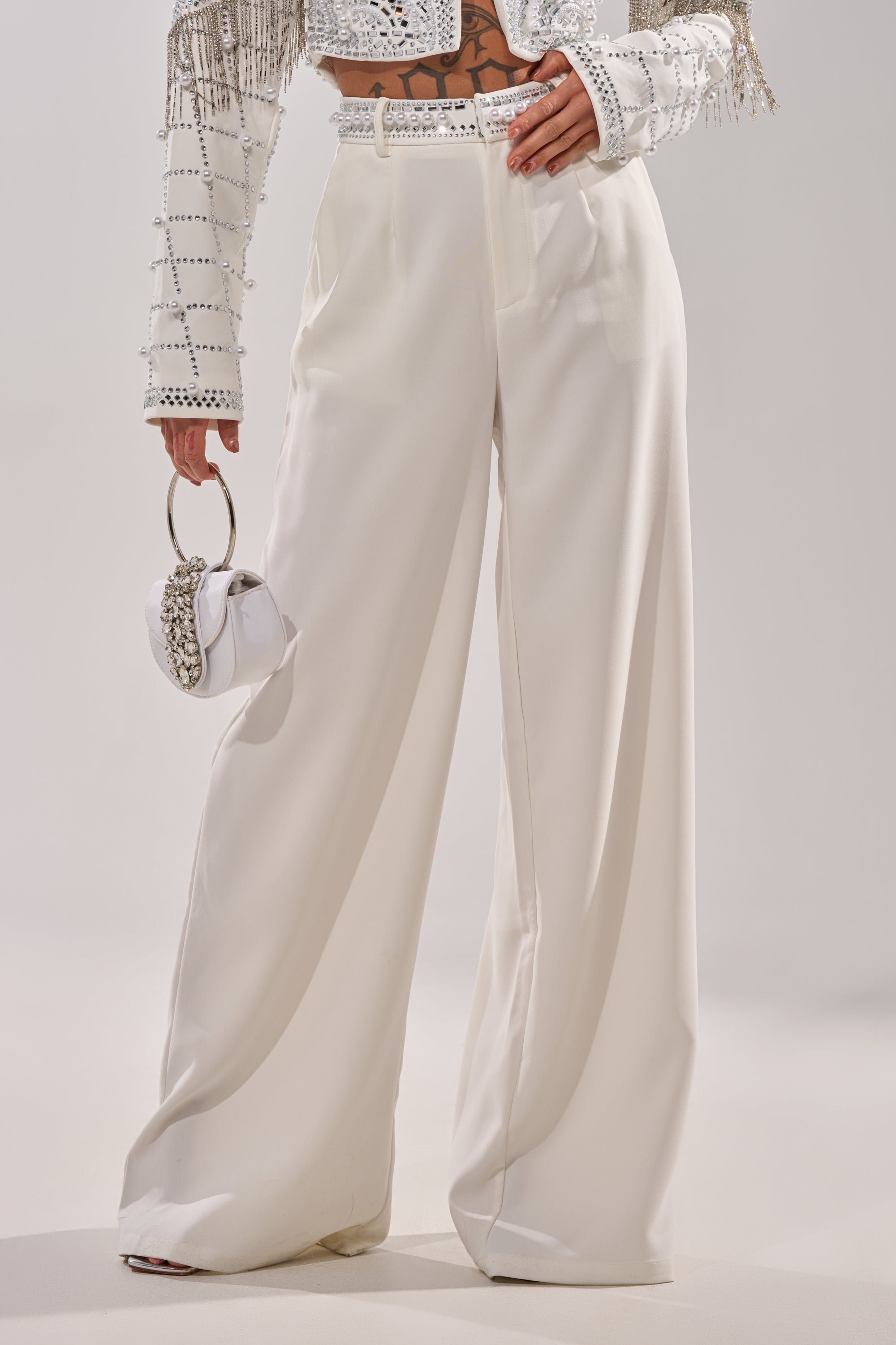 Wearing DIRTY MARTINI EMBELLISHED TROUSER IN WHITE—high-waisted, wide-leg, high-rise—paired with a white embellished cropped jacket and a small white circular-handle handbag. The plain background is light-colored. 2