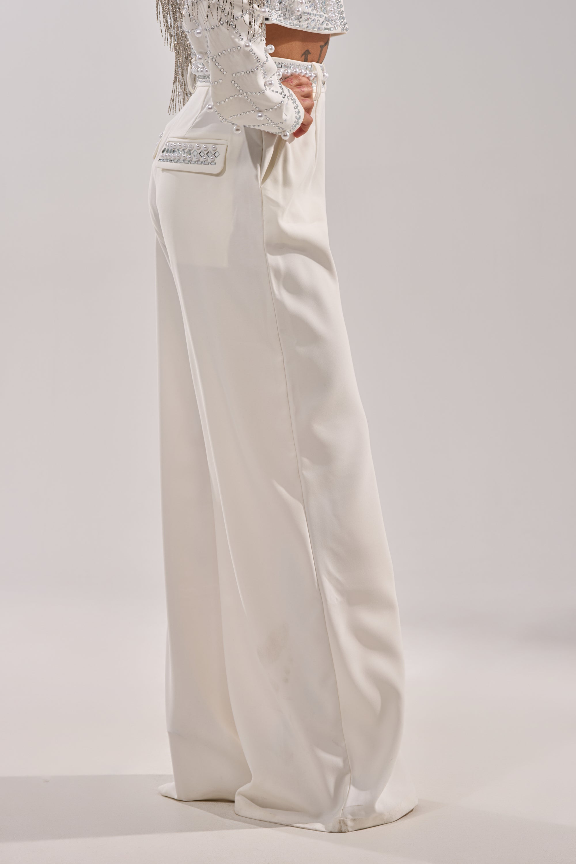 A person stands against a plain background wearing the DIRTY MARTINI EMBELLISHED TROUSER IN WHITE, featuring high-waisted, wide-leg styling and decorative back pocket stitching, paired with a cropped embellished top to highlight the chic fit. 3