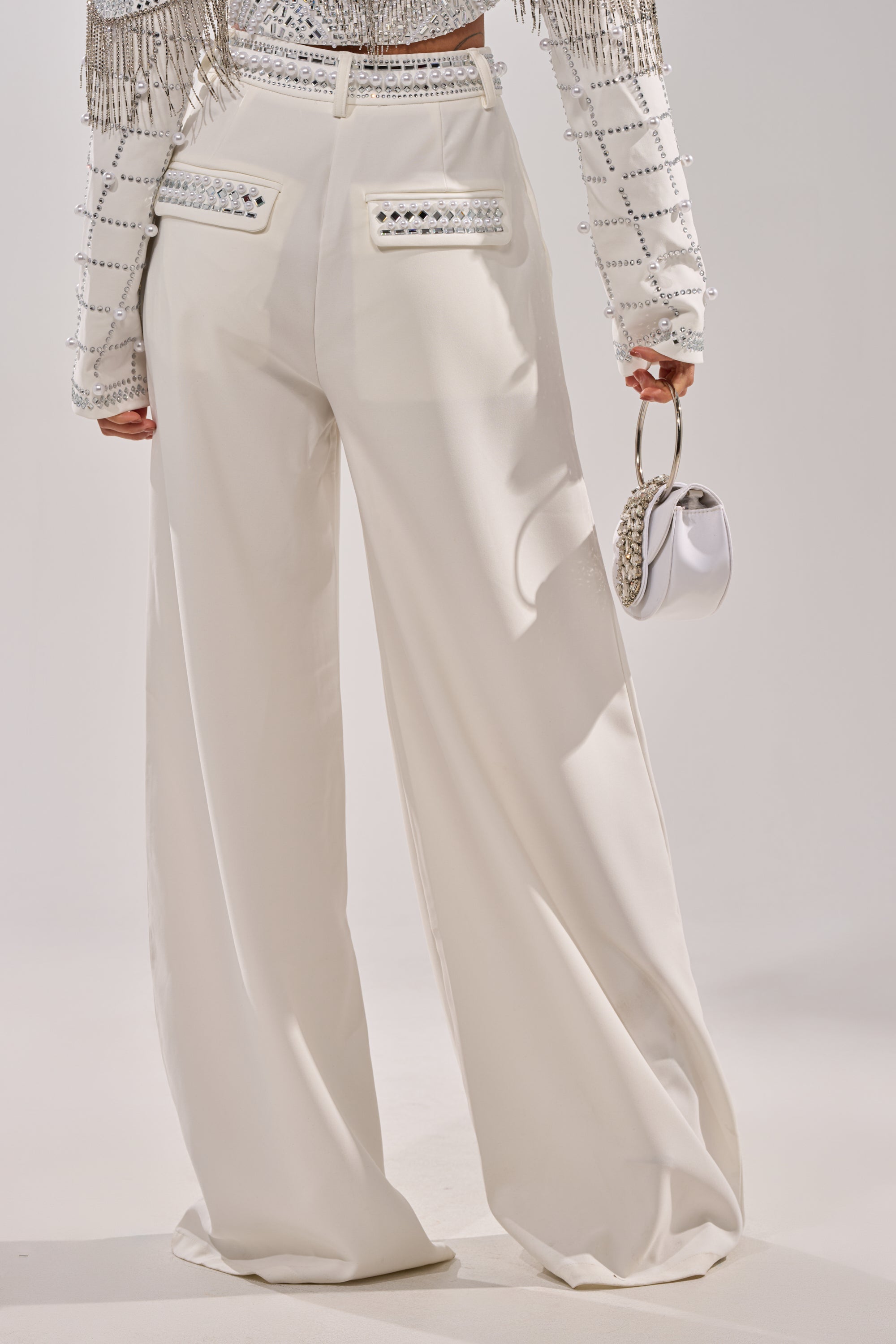 Wearing the DIRTY MARTINI EMBELLISHED TROUSER IN WHITE—high-waisted, wide-leg pants with ruched details, studded pockets—paired with a sparkling fringe-beaded top and a small white handbag. 4
