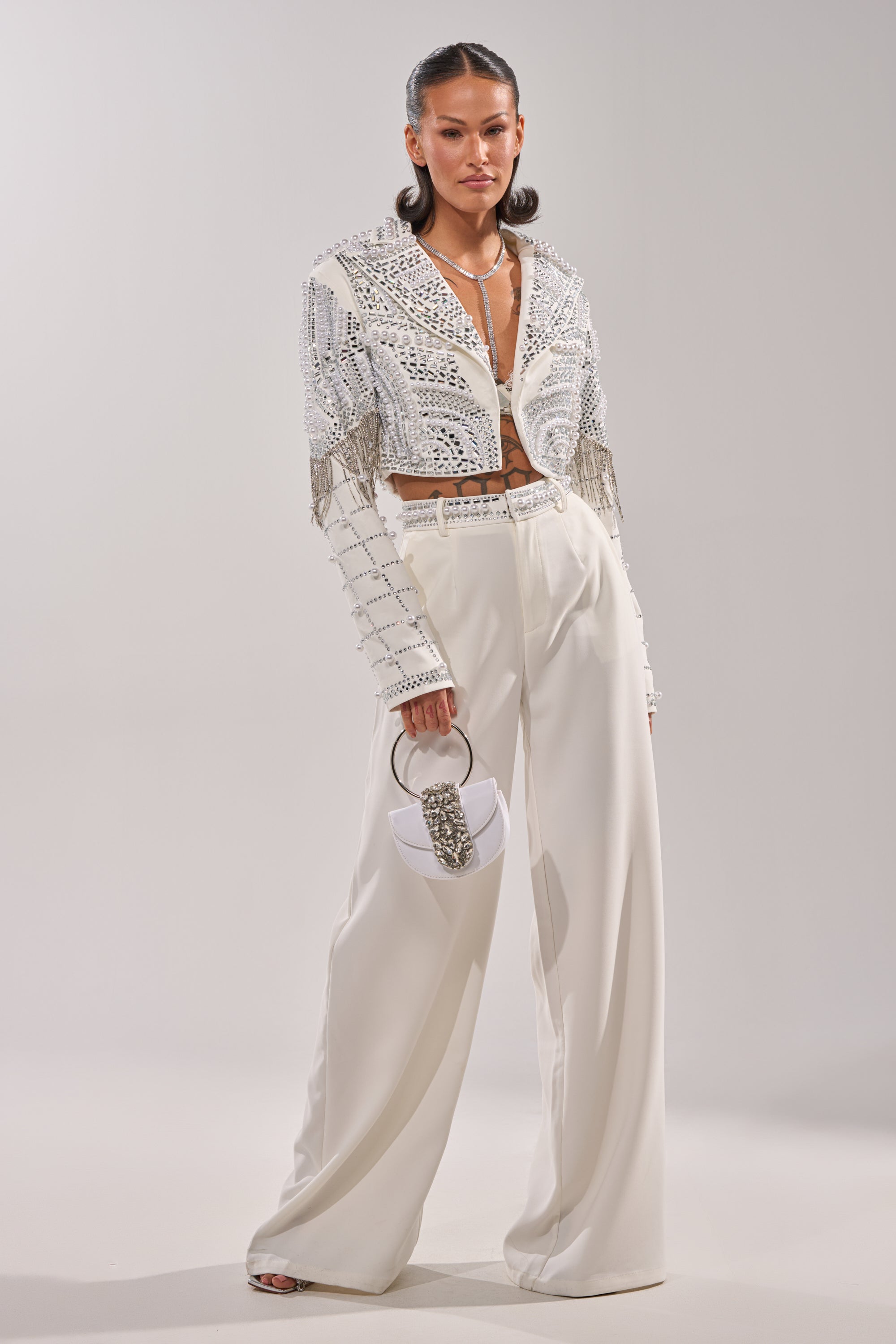 A woman stands confidently in the DIRTY MARTINI EMBELLISHED TROUSER IN WHITE, paired with a cropped embellished jacket, sheer top, silver heels, and a small white handbag. 5