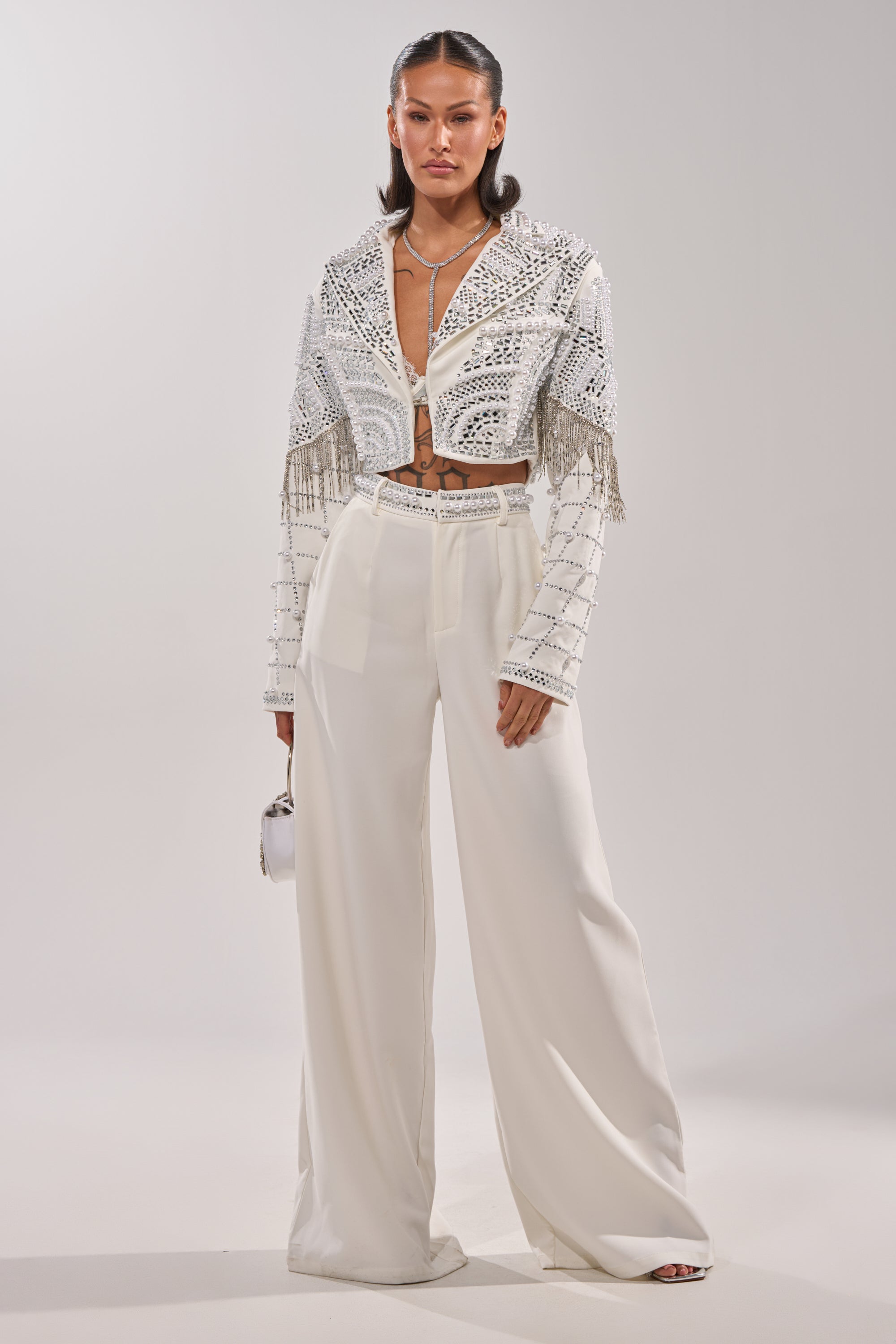 A woman stands against a plain background wearing DIRTY MARTINI EMBELLISHED TROUSER IN WHITE, paired with a matching crop top and cropped jacket with silver embellishments and fringe, accessorized with a white handbag and layered necklaces. 6