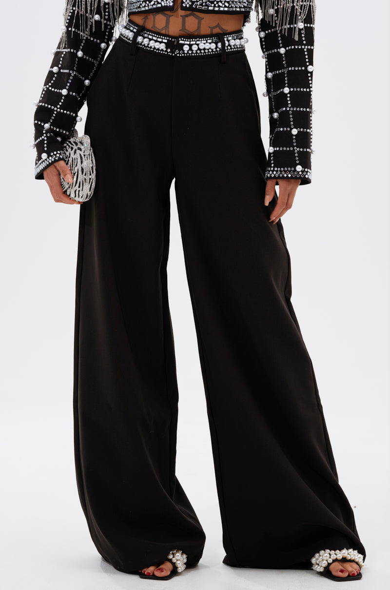 A person wearing DIRTY MARTINI EMBELLISHED TROUSER, a cropped geometric-patterned beaded fringe jacket, holding a textured clutch, and pearl-embellished open-toe shoes.
