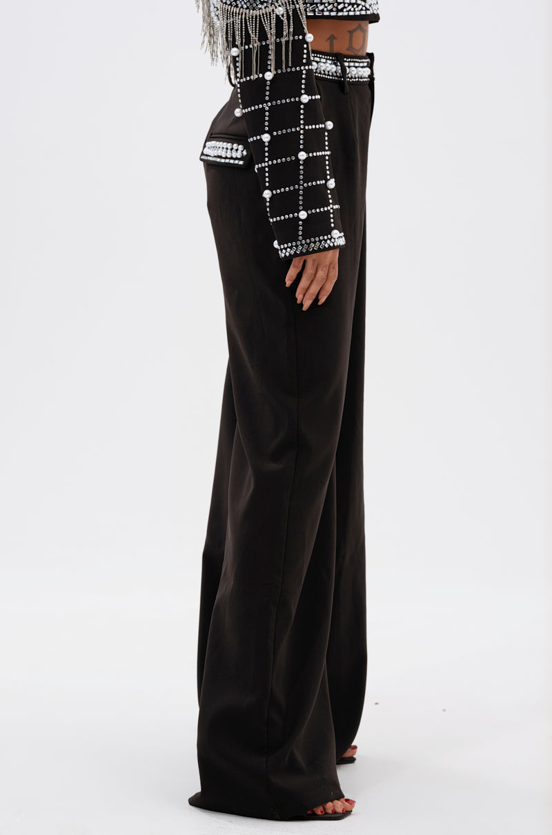 A person stands sideways wearing the DIRTY MARTINI EMBELLISHED TROUSER, a high-rise wide-leg black pant with a pearl rhinestone waistband, paired with a black cropped jacket with white grid patterns, silver studs, and fringe. The background is plain white.