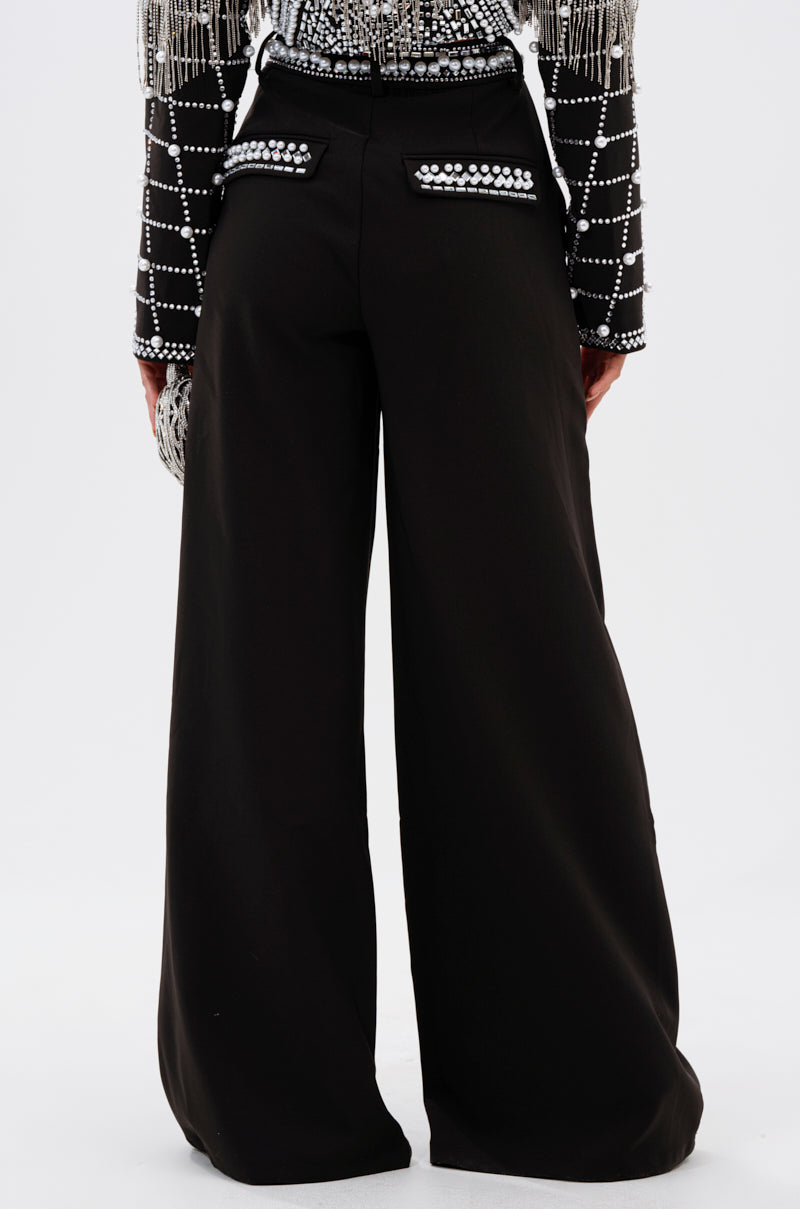A person is shown from the back wearing the DIRTY MARTINI EMBELLISHED TROUSER, featuring high-rise wide legs and silver-studded pockets, paired with a black top adorned with silver beads in grid patterns, against a plain background.